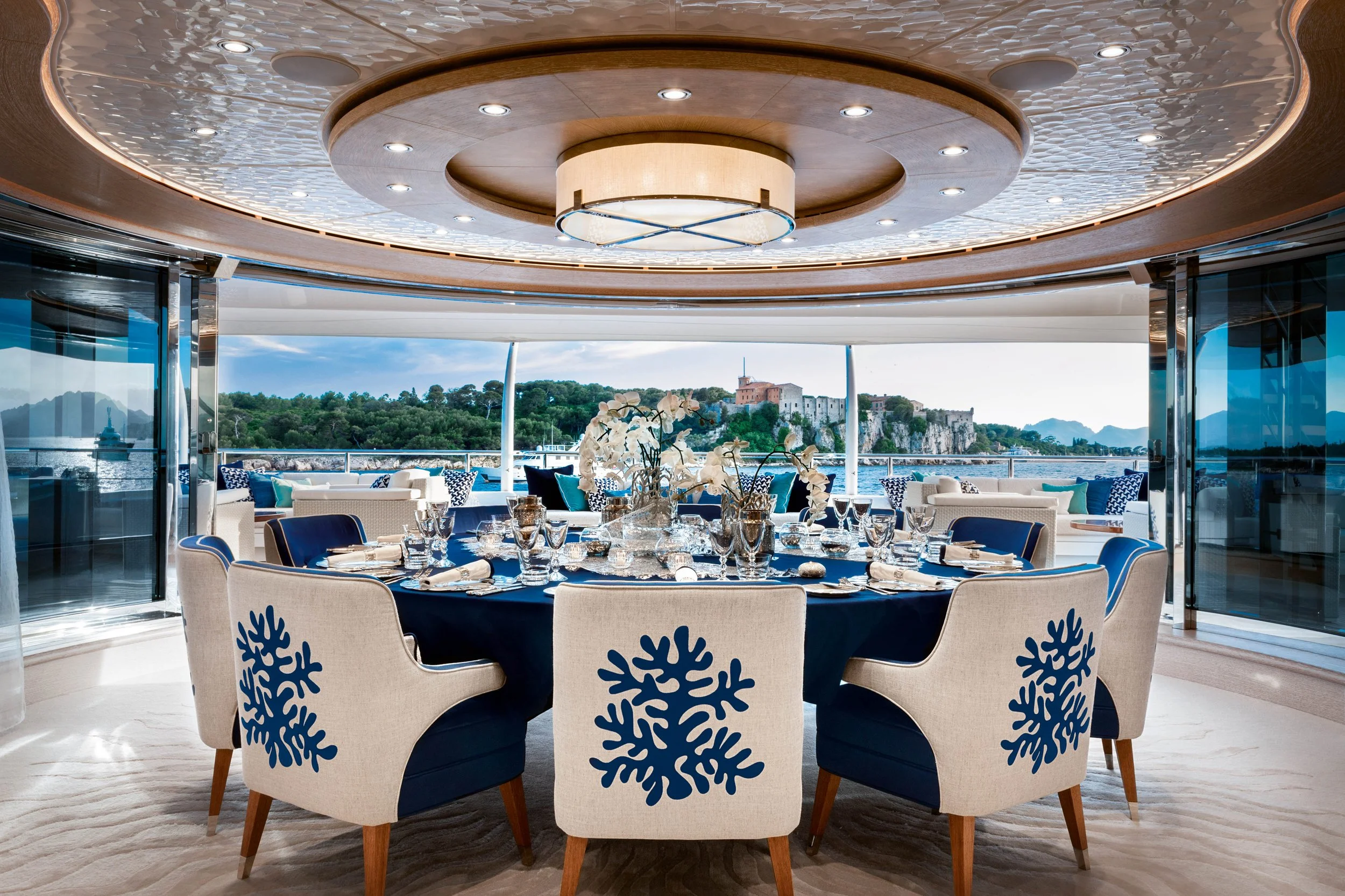 A luxurious dining area on a yacht with a round table set with glassware, surrounded by chairs with blue snowflake patterns. The space has a curved ceiling with built-in lights, and large sliding glass doors that open to an outdoor view of a castle on a hill, water, and greenery.