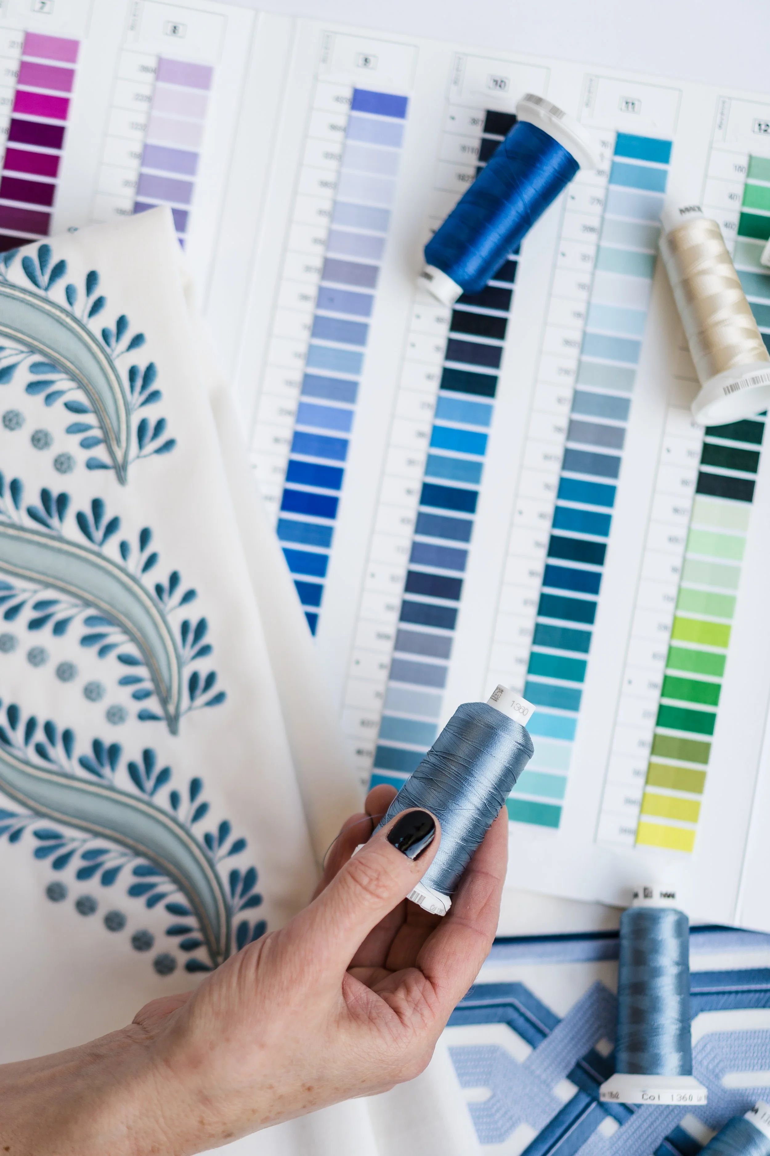 A person's hand holding a spool of gray embroidery thread, with fabric displaying blue embroidered textile designs, color charts, and additional spools of thread in the background.