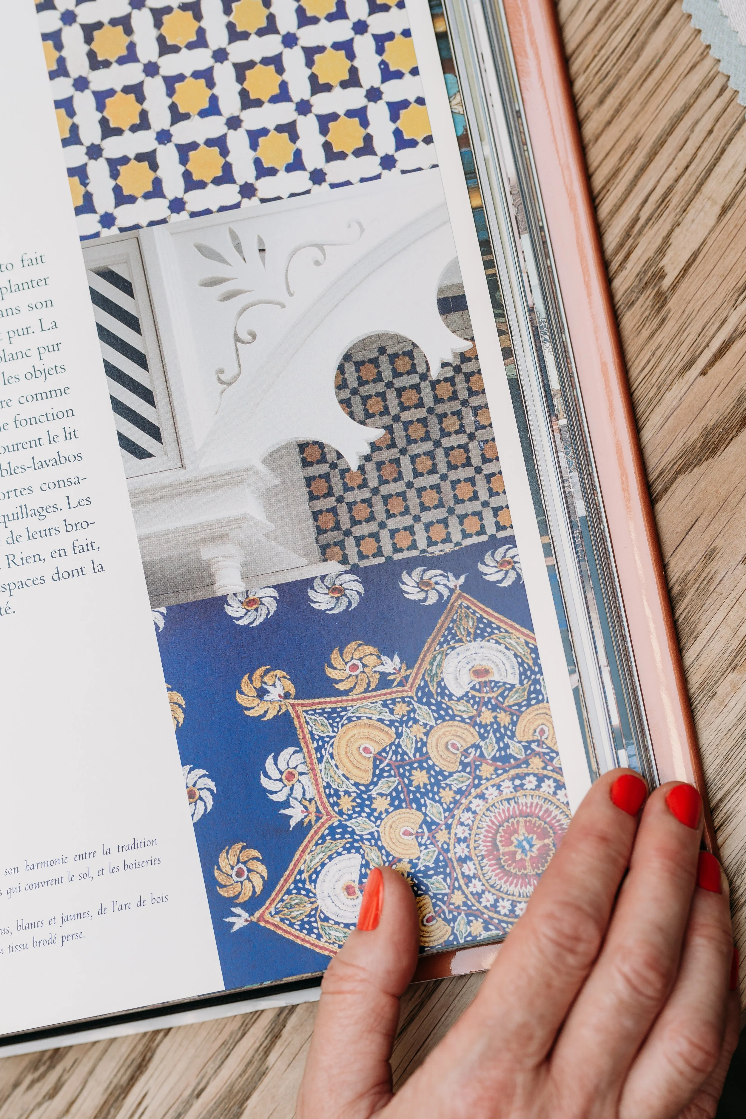Close-up of a person with red-painted nails holding a book open to a page showing images of intricate tile and carpet designs with various patterns and colors.