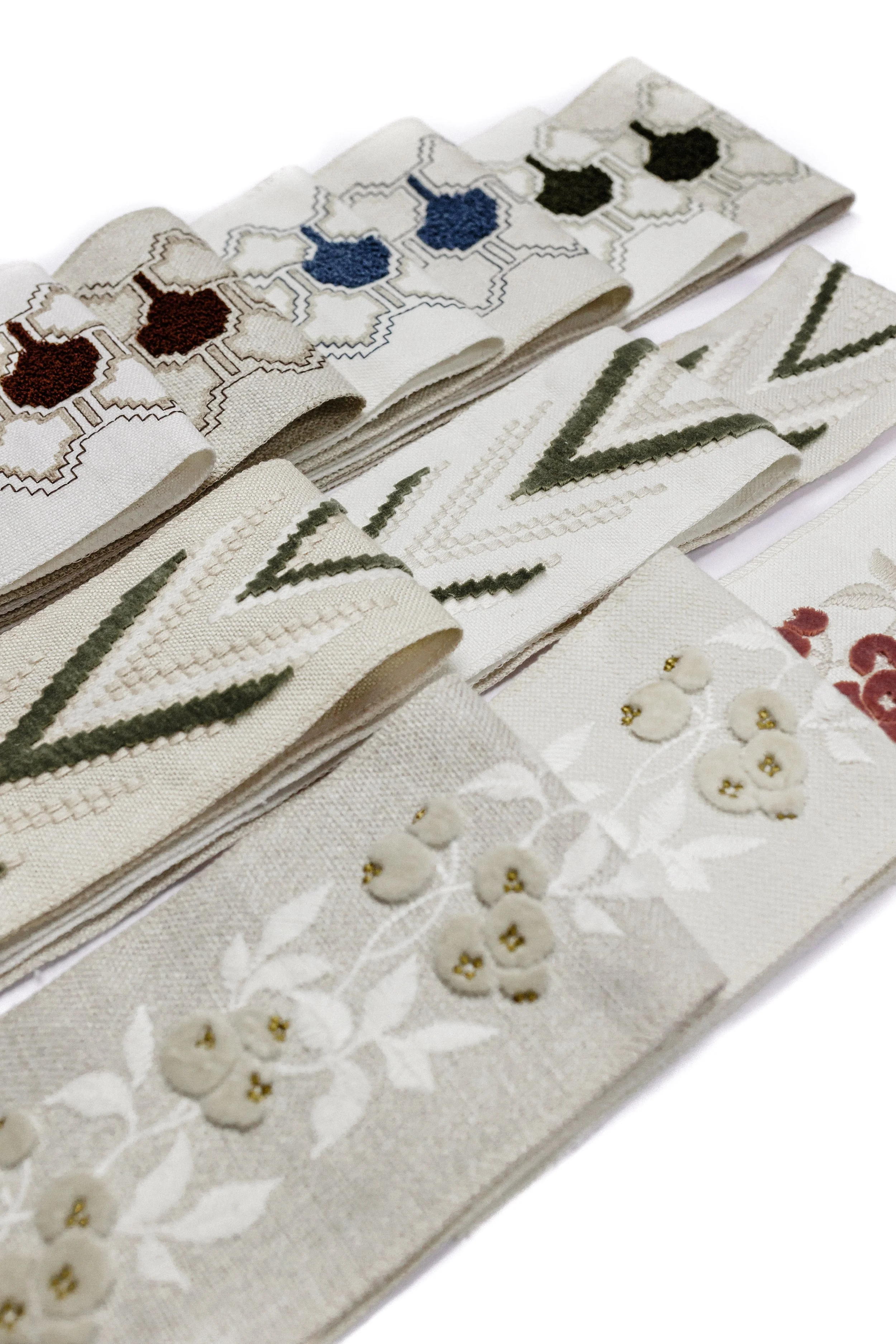 Close-up of folded white linen napkins with embroidered floral and geometric designs in various colors, including brown, blue, green, and white, displayed on a white background.
