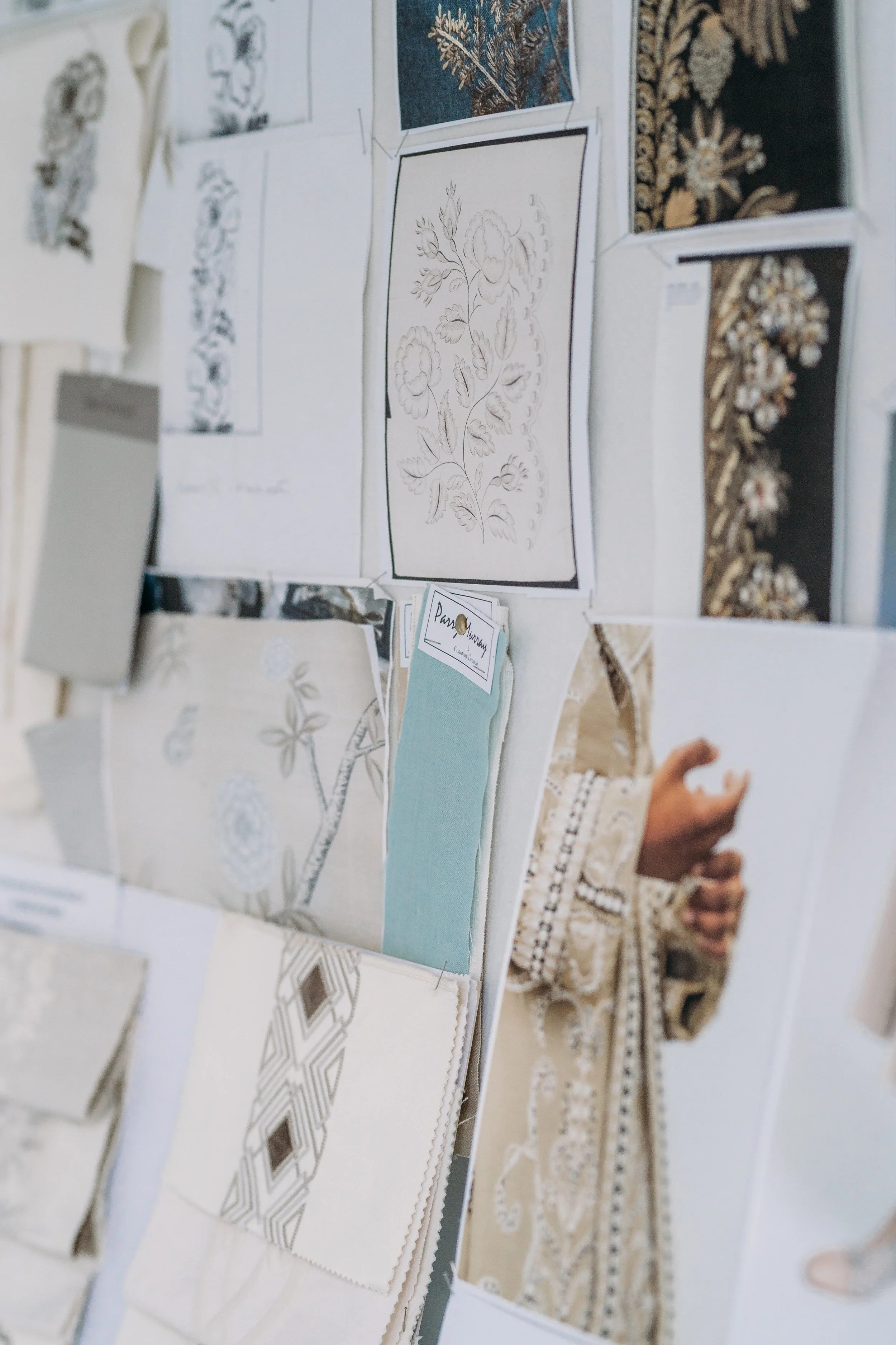 A display of various fabric samples and embroidery patterns, some with floral designs, on a wall or board. Part of a person's hand and arm are visible, with the person wearing long sleeves, in the background.