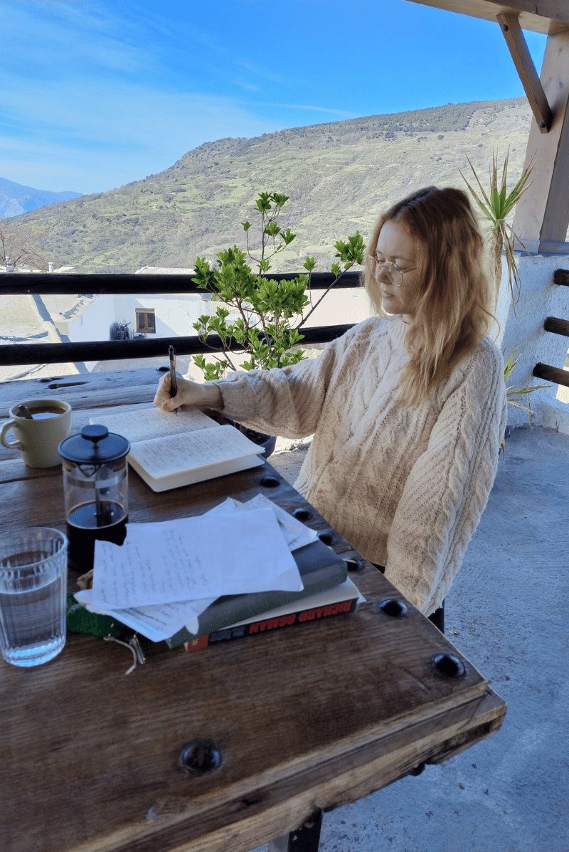 Author Robin Brookes, wearing glasses and a white knitted sweater, sits at a wooden table on a balcony, writing in a notebook. She is surrounded by a cup of coffee, a French press, water glasses, and scattered papers.