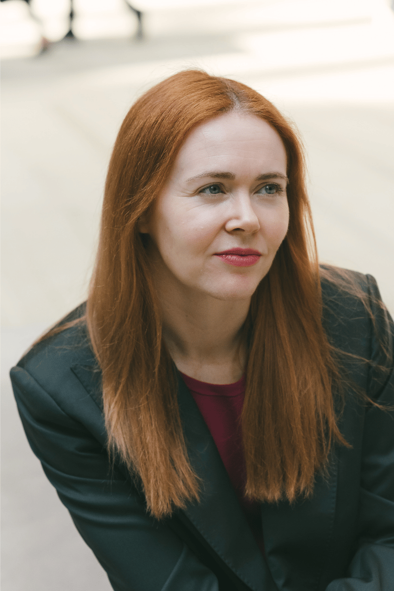 Author and playwright Robin Brookes with long red hair, wearing a dark blazer and a maroon shirt, sitting outdoors.
