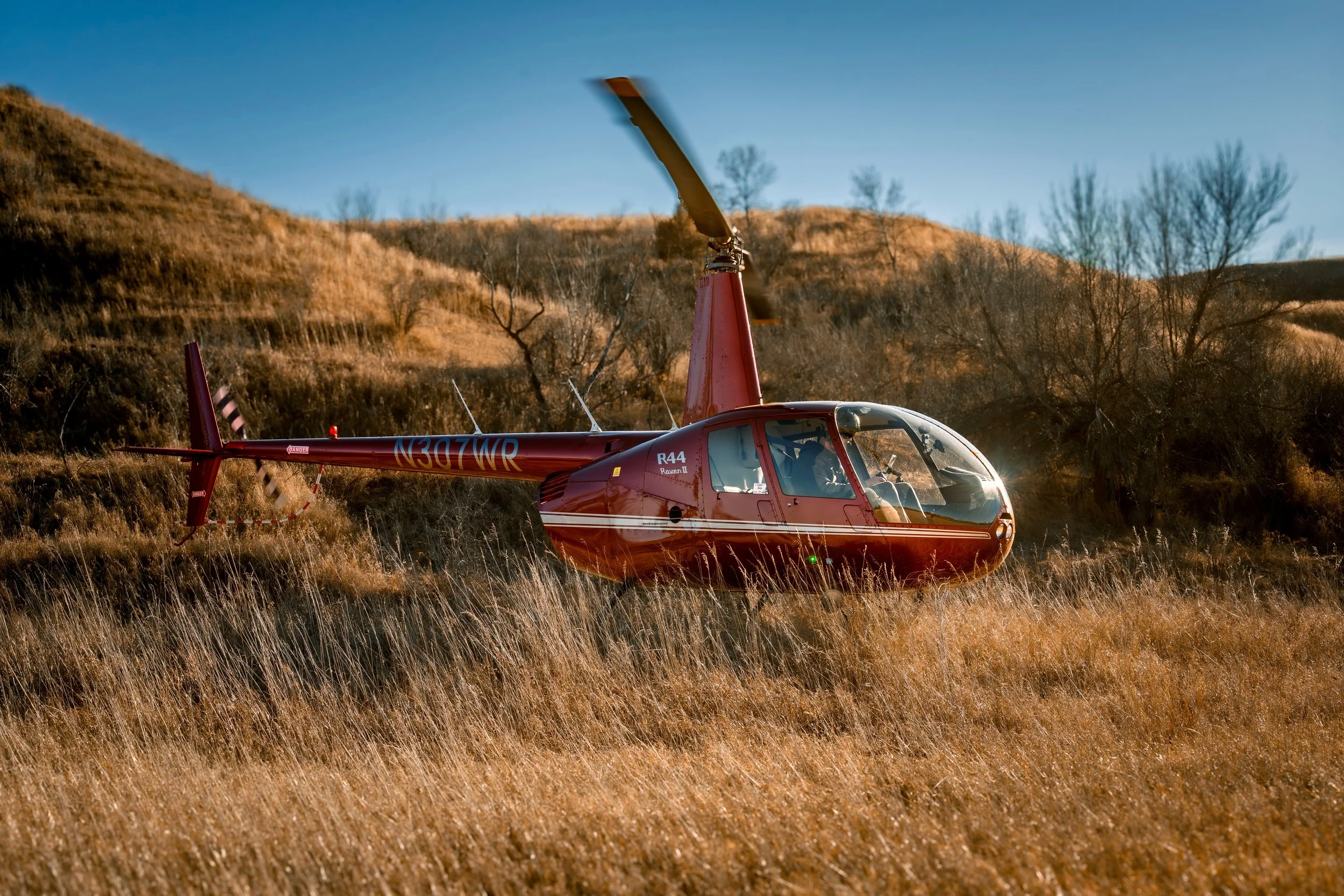 Red helicopter on a grassy hillside with trees and dry hills in the background, under a blue sky.