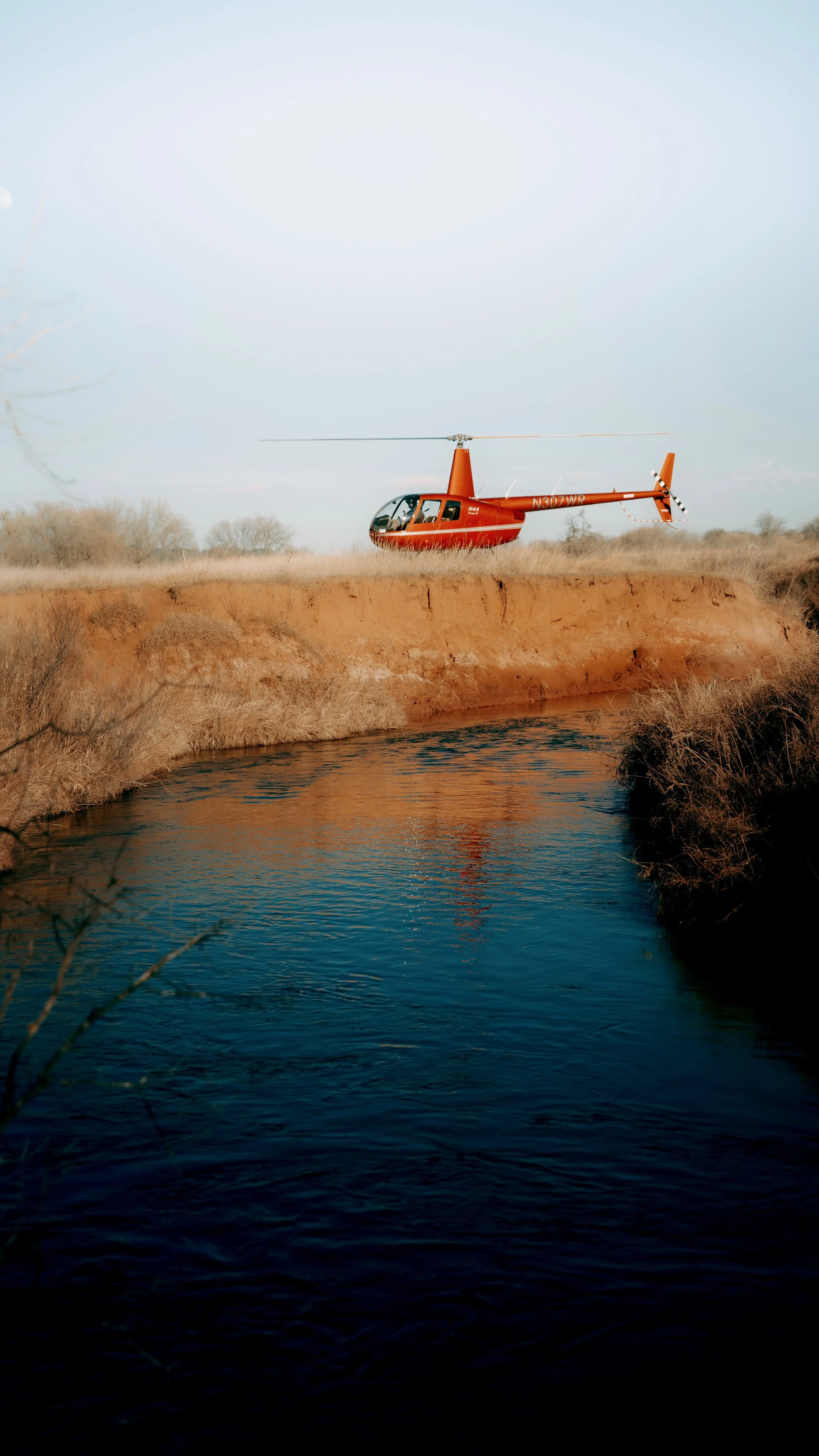 A red helicopter appears to be parked near a river with trees in the background and clear sky.