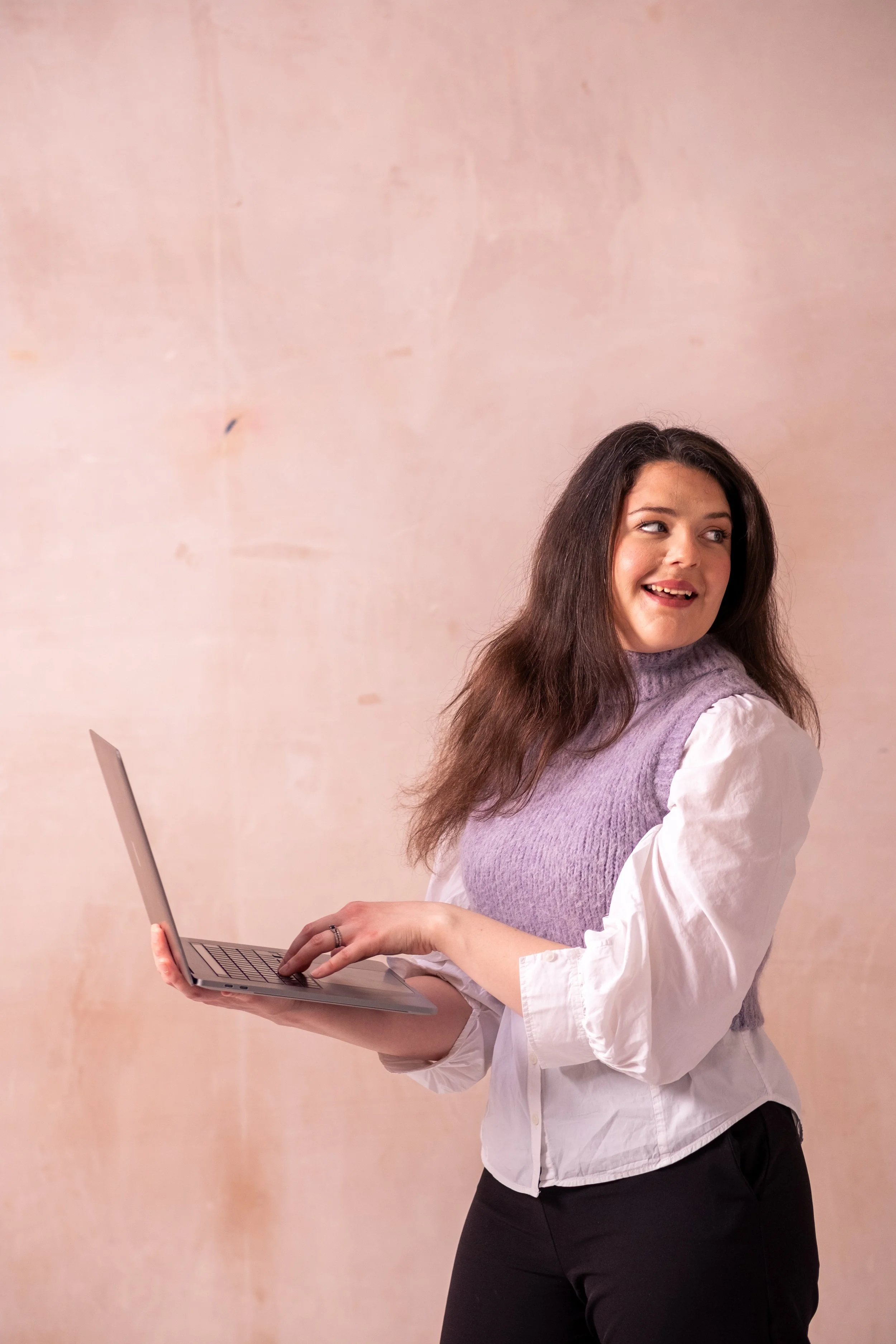 Bethany Stone, wearing a white shirt and lilac sweater vest, body facing the left of the screen looking to the right over her shoulder. She holds a laptop and stands against a neutral background.