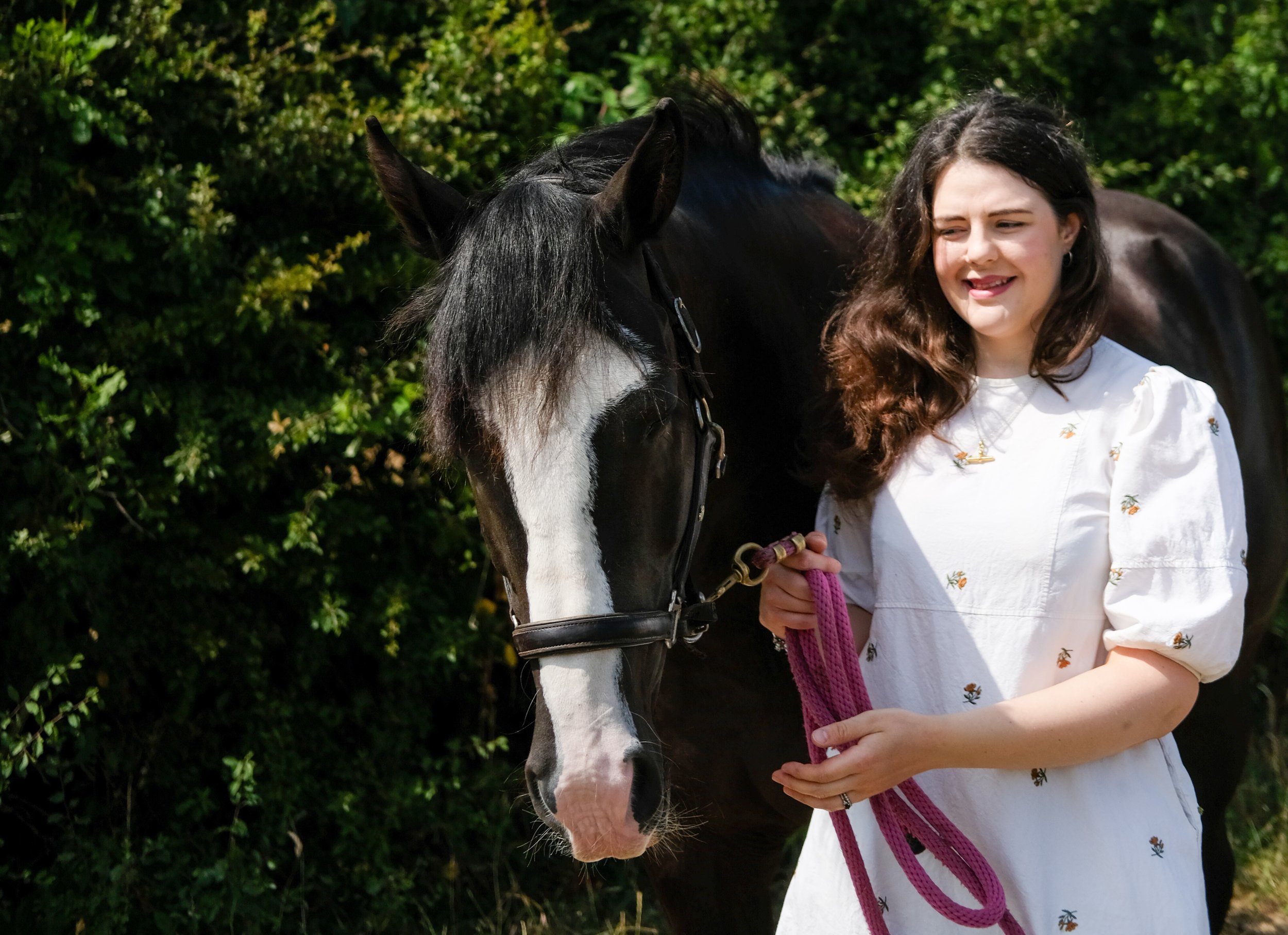 Bethany Stone stood facing the camera holding a pink leadrope attached to the leather headcollar of a dark bay horse with a white blaze
