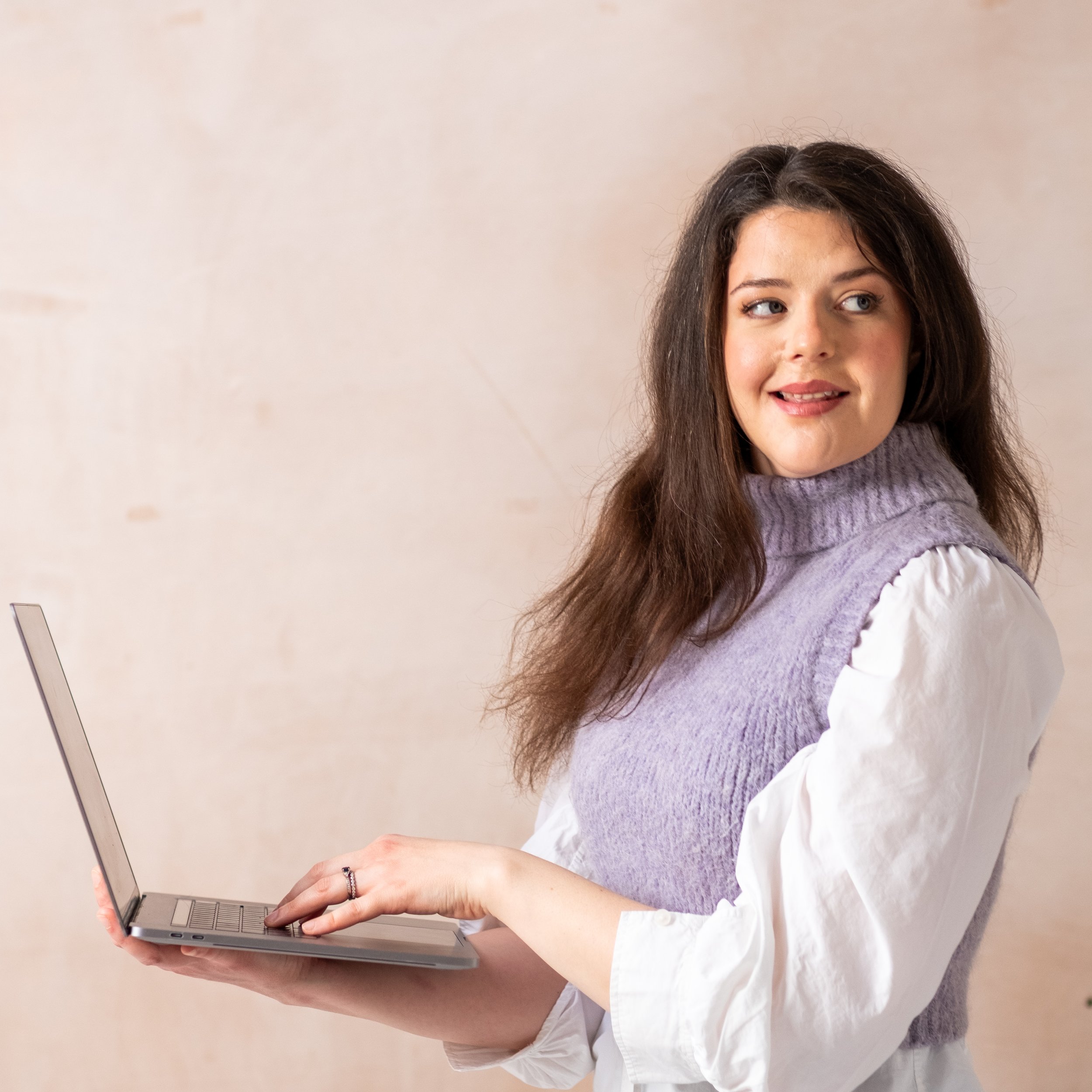 Bethany Stone stands in profile holding a laptop. She looks her her left shoulder, her left hand is poised over the keyboard
