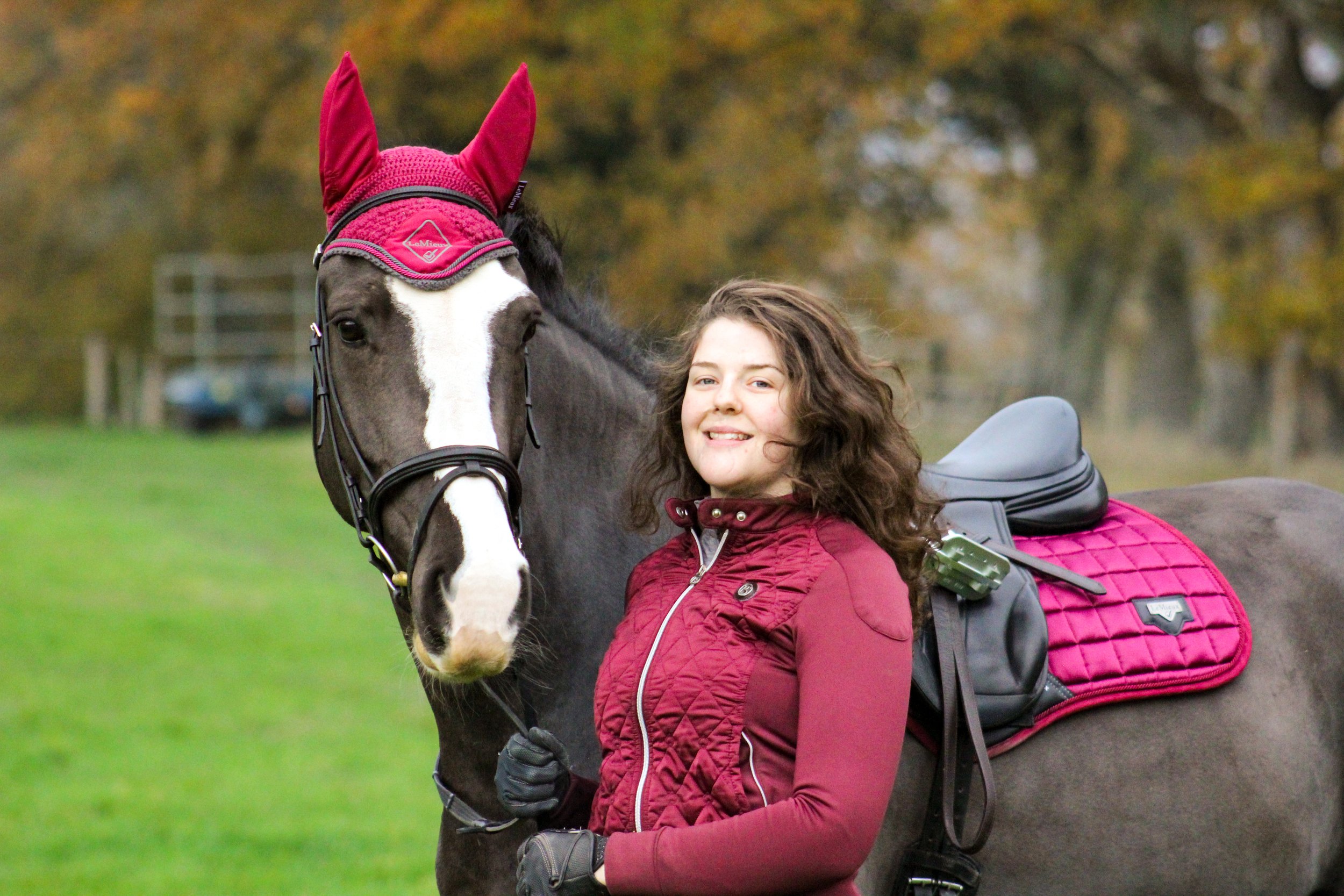 Close up of Bethany Stone with a dark bay horse who has a white blaze in an autumnal field. The horse wears a saddle and bridle with a rose coloured ear bonnet and saddle pad. Bethany wears a matching quilted jacket.