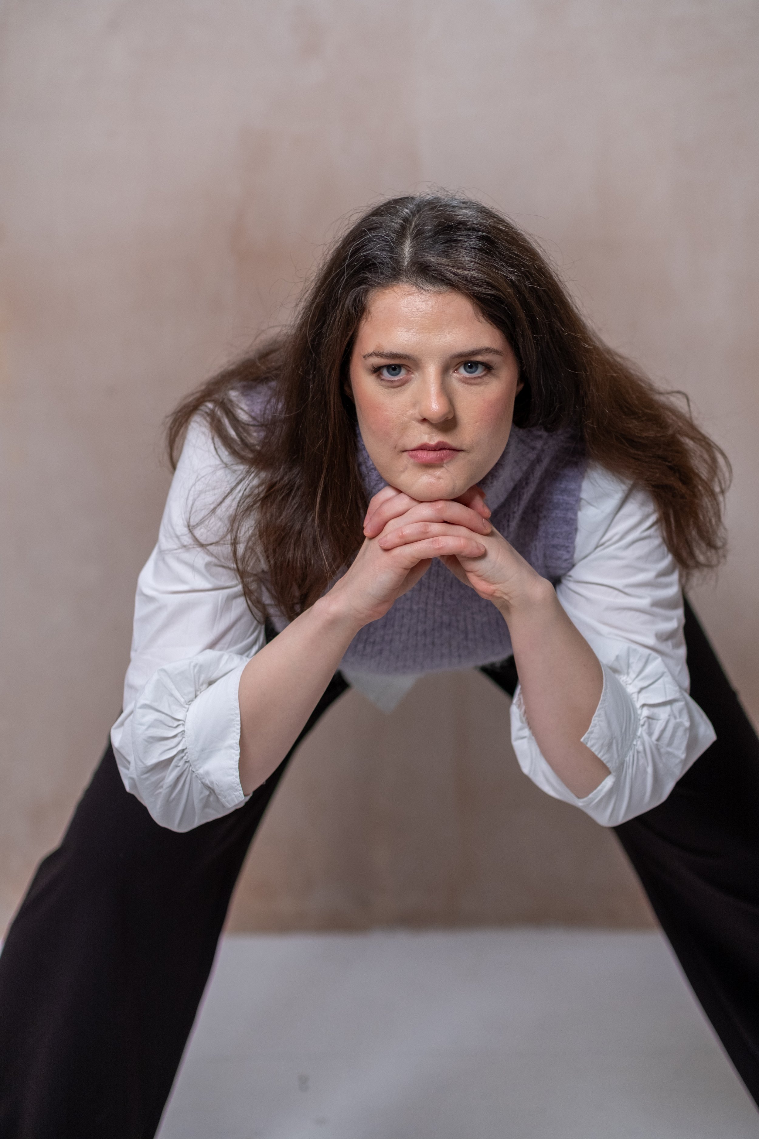 Bethany Stone leans forward with a wide leg stance, hands clasped under her chin with a serious face. She is copying an iconic pose by actor Willam Dafoe.