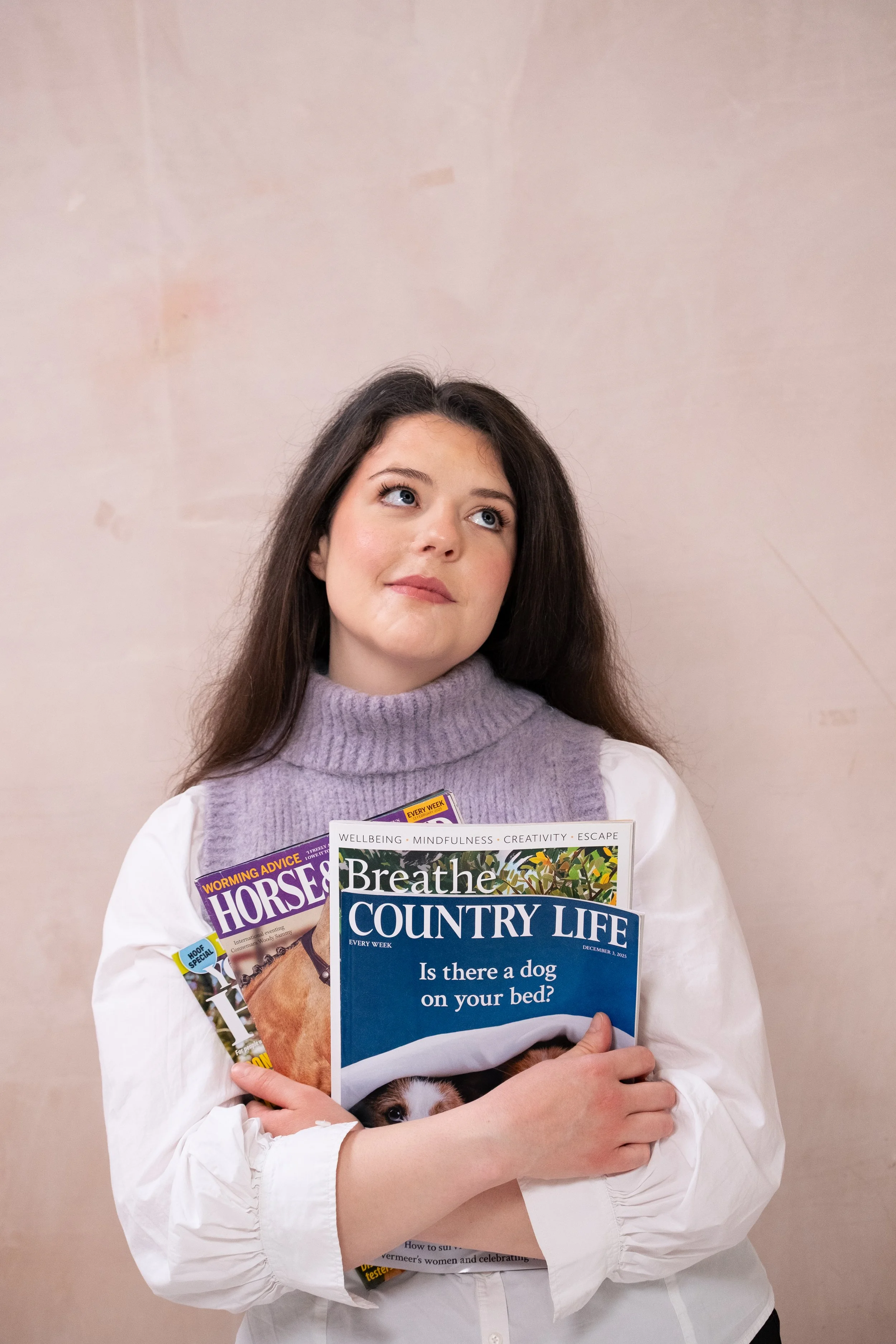 Close up of Bethany Stone who wears a white shirt and a lilac sweater vest. She looks up and to the right. In her arms she holds four magazines.: Country Life, Breathe, Horse & Hound and Your Horse.