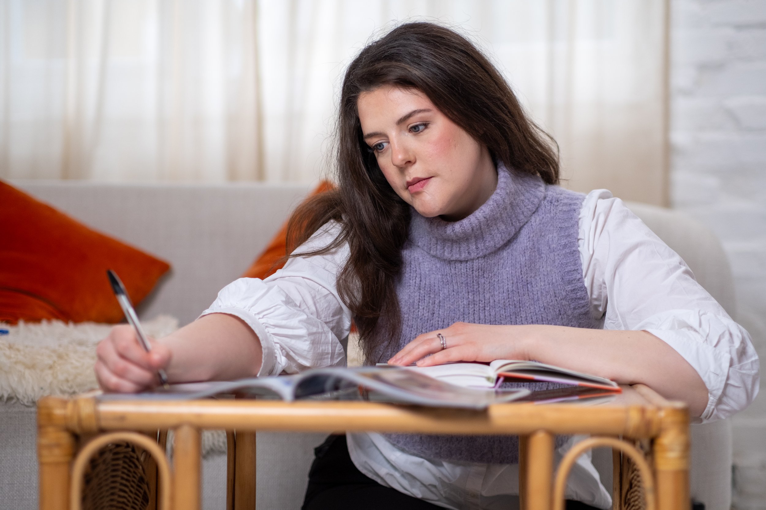 Bethany Stone kneels on the floor and writes on a notebook, leaning on a class coffee table. A grey sofa with burnt orange cushions is behind.