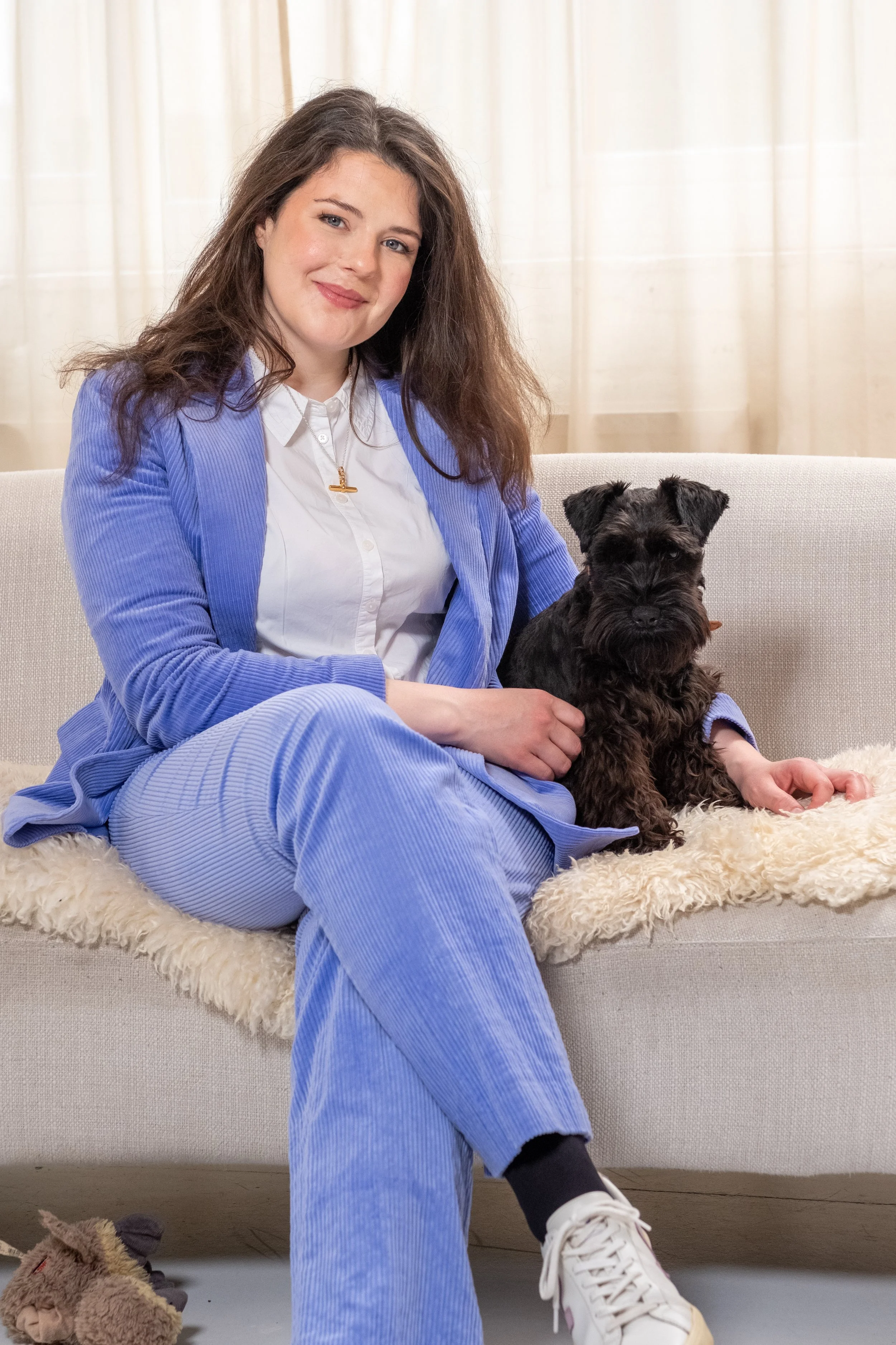 Bethany Stone sitting on a beige sofa. Beneath her is a sheepskin style rug. She has her arm around a dog, a black miniature schnauzer, who sits next to her. She is wearing a lilac corduroy suit with a white shirt and white trainers.