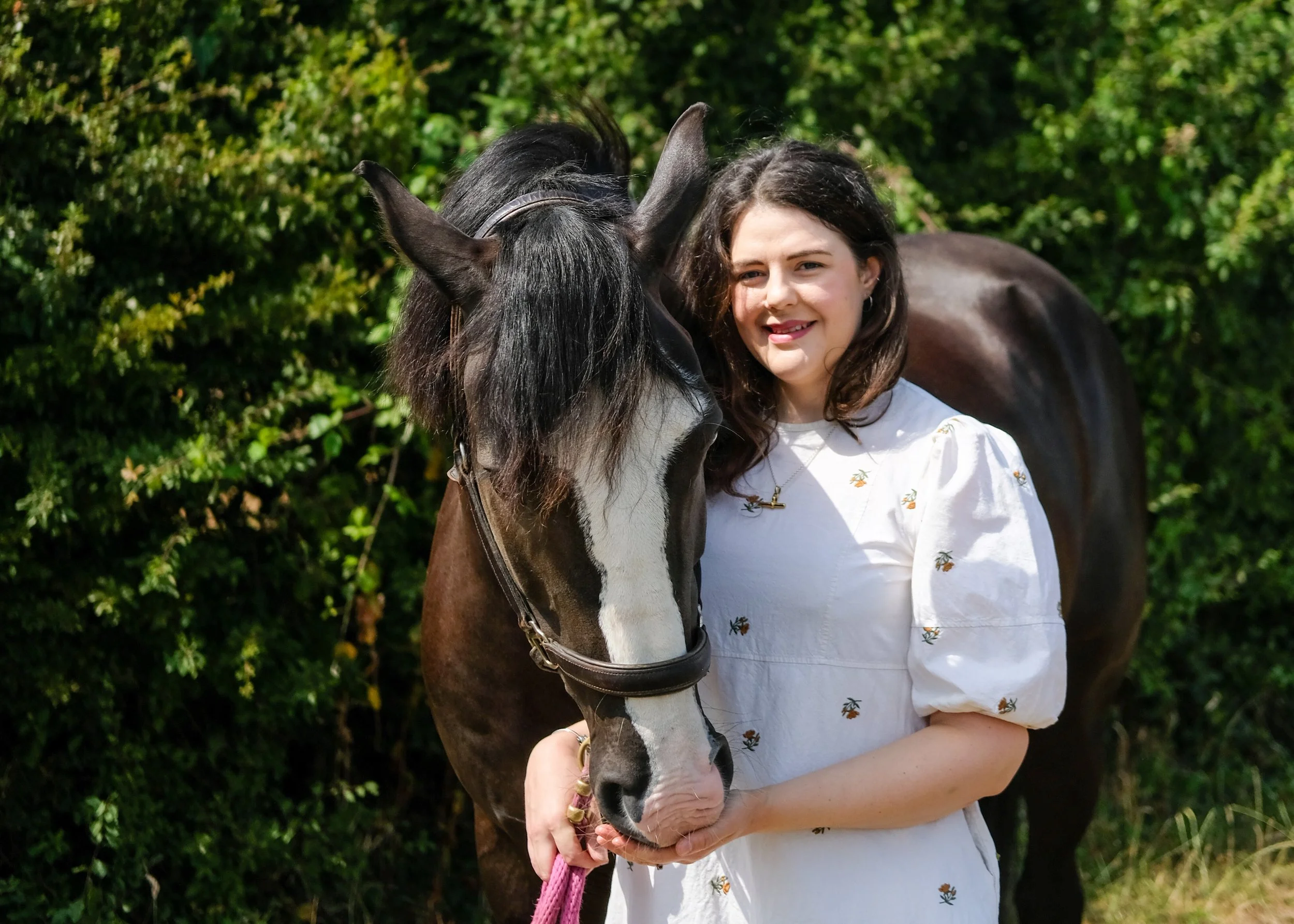 Bethany Stone stood with a dark bay horse in front of a hedge. The horse’s head is over her right shoulder. They are stood facing the camera. Bethany wears a beige dress with floral details.