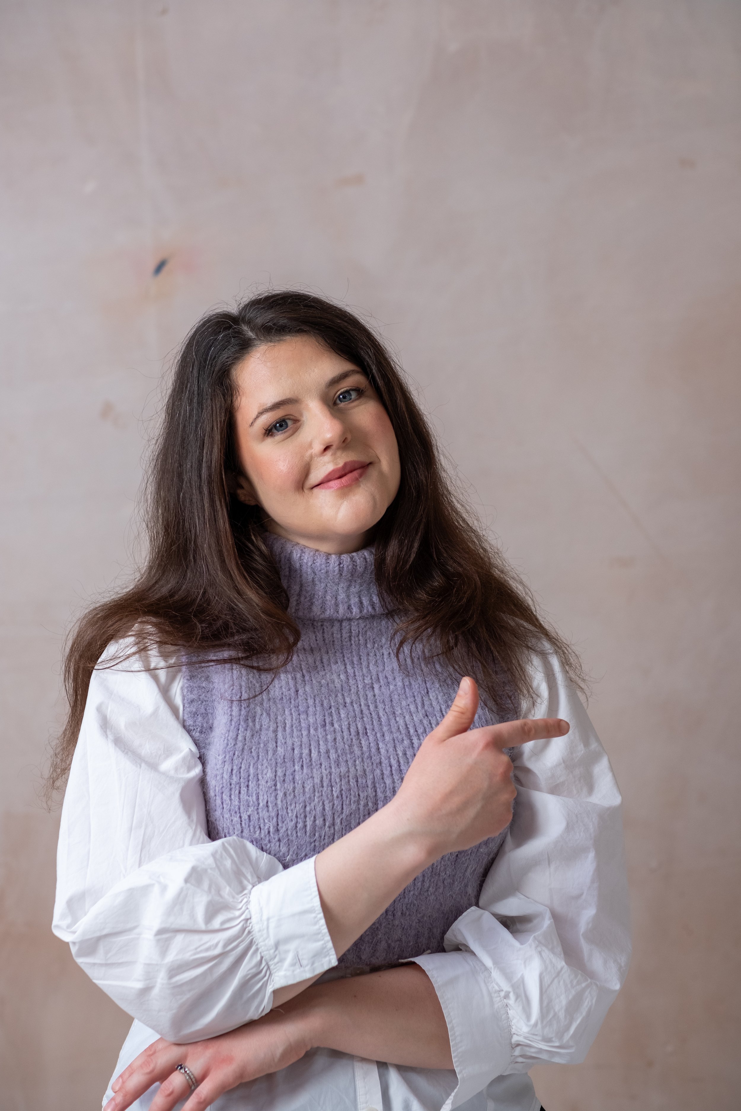 Bethany Stone, a woman with long brown hair, stood against a neutral background. She is wearing a white shit with large sleeves and a lilac high neck sweater vest. She is looking at the camera and and is pointing to the right of the screen