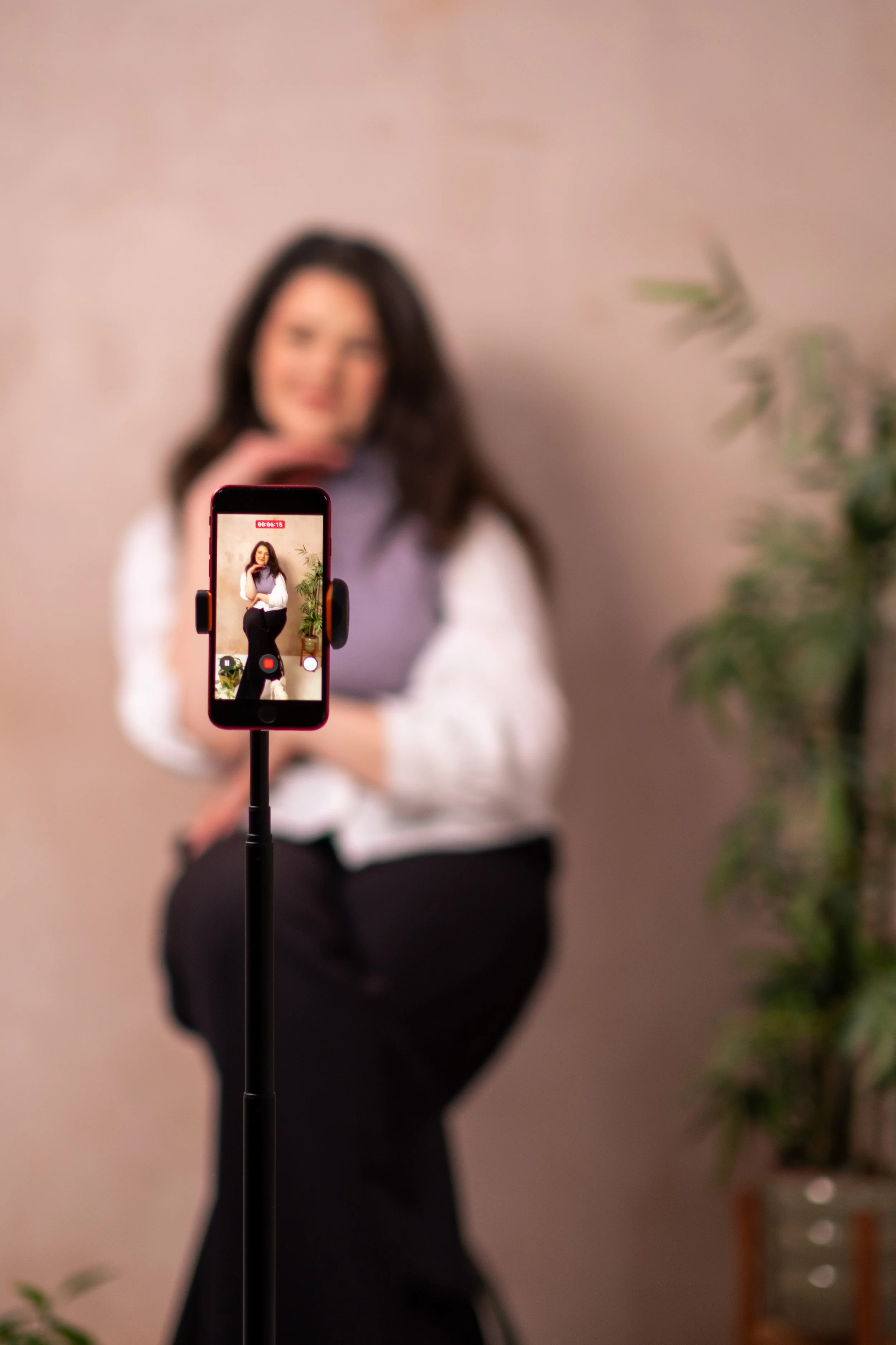 An out-of-focus-image of Bethany Stone sat on a stool, wearing a white shirt, lilac sweater vest and black trousers. In the foreground is a tripod with an iPhone filming her