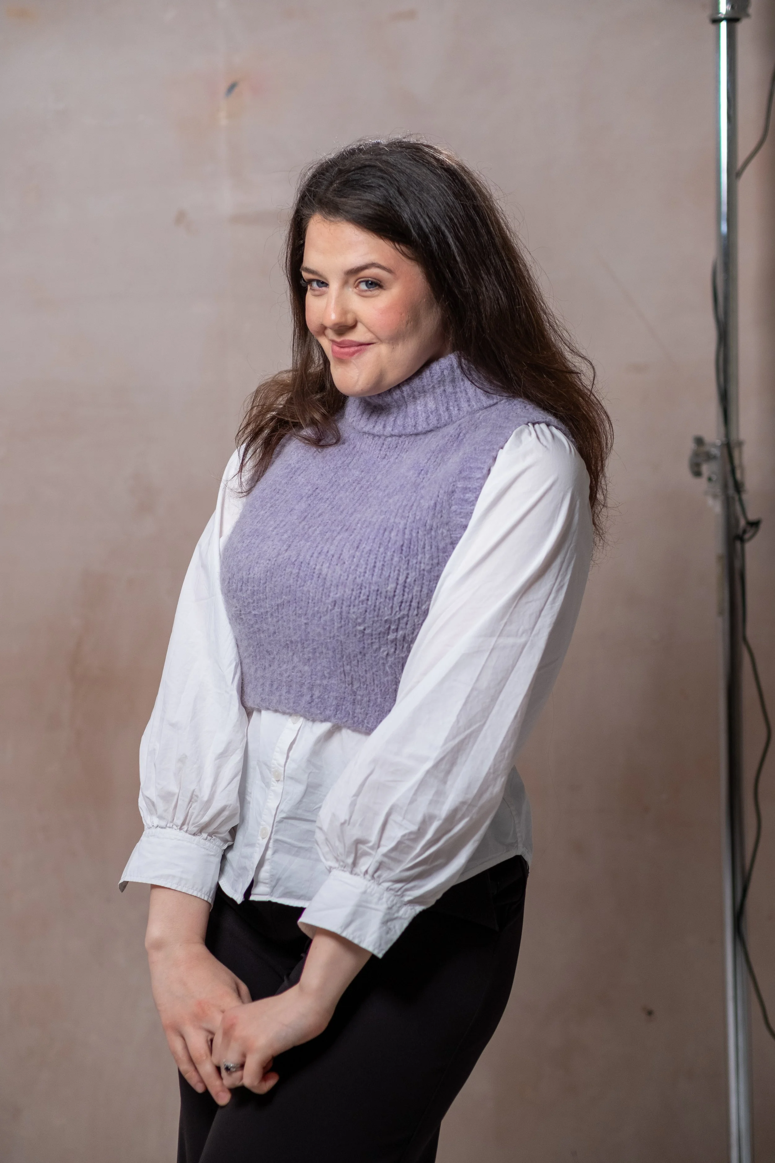 Bethany Stone stands against a neutral background wearing a white shirt, lilac sweater vest and black trousers. Her hands are clasped together and she smiles cheekily.