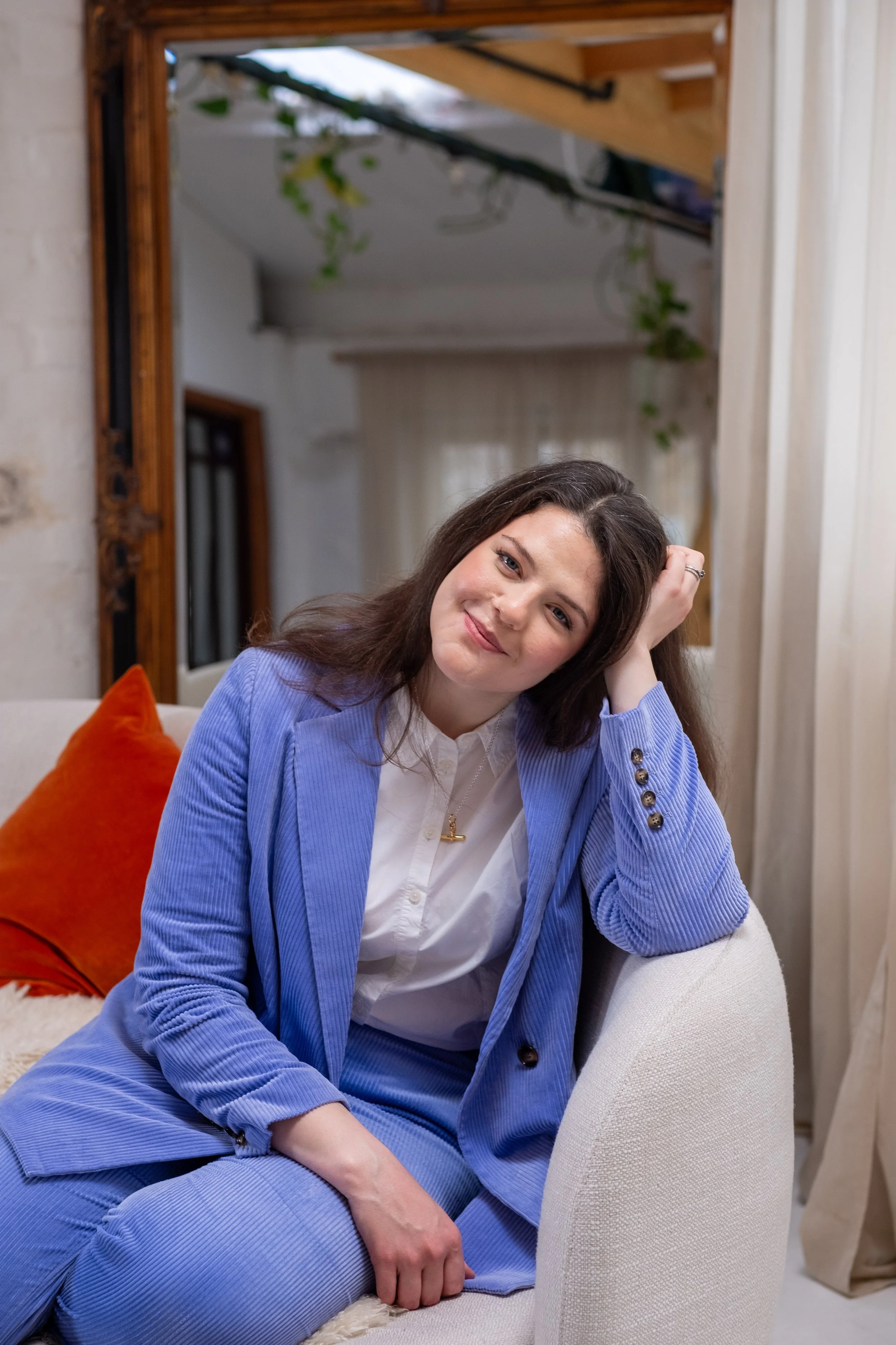 Bethany Stone wearing a two-piece lilac suit and white shirt. She sits facing the camera on the sofa with her left elbow resting on the back and her head resting on her left hand.