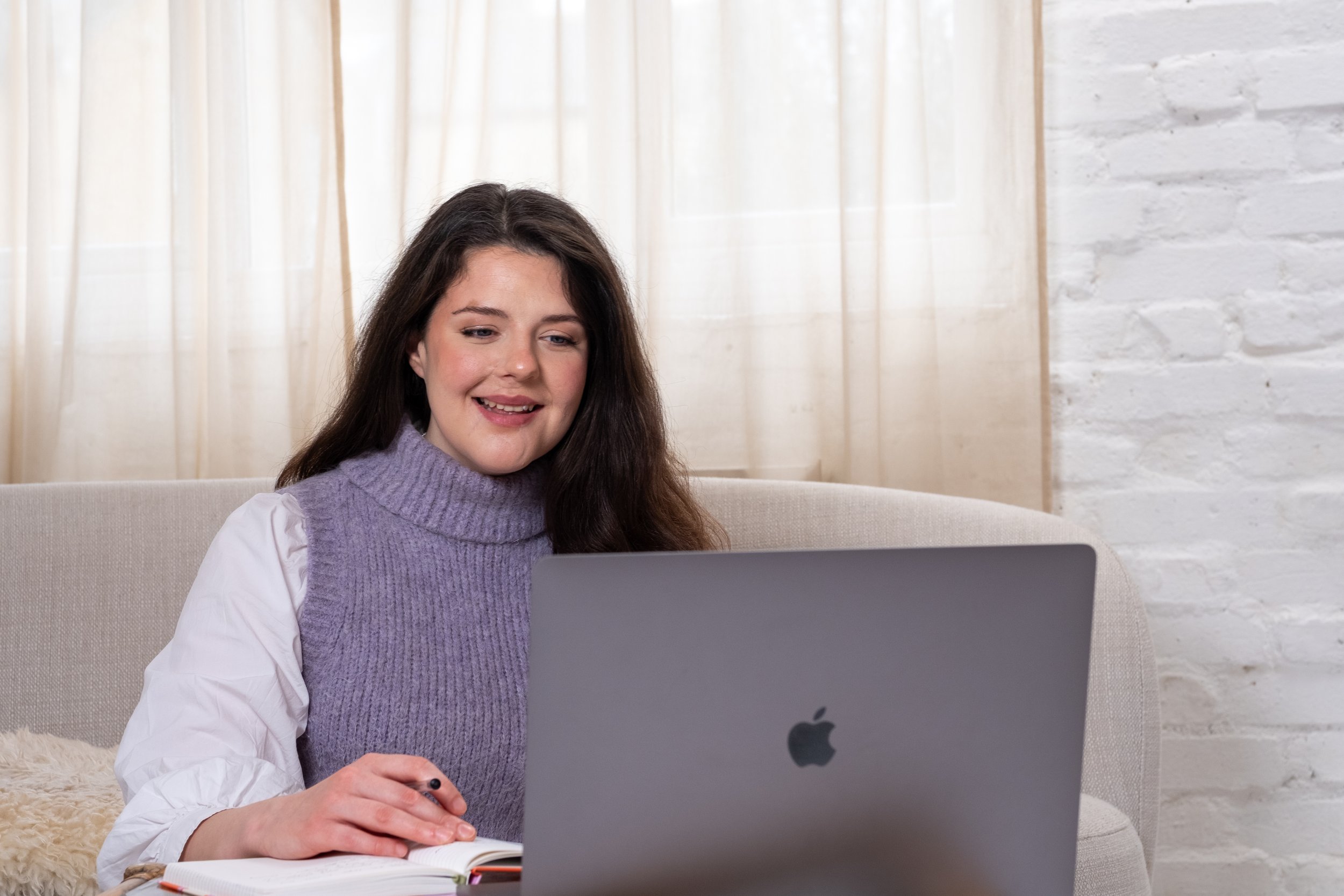 Bethany Stone sits in front of Macbook, looking at the screen and smiling. In her right hand is a pen, under her hand is a book.