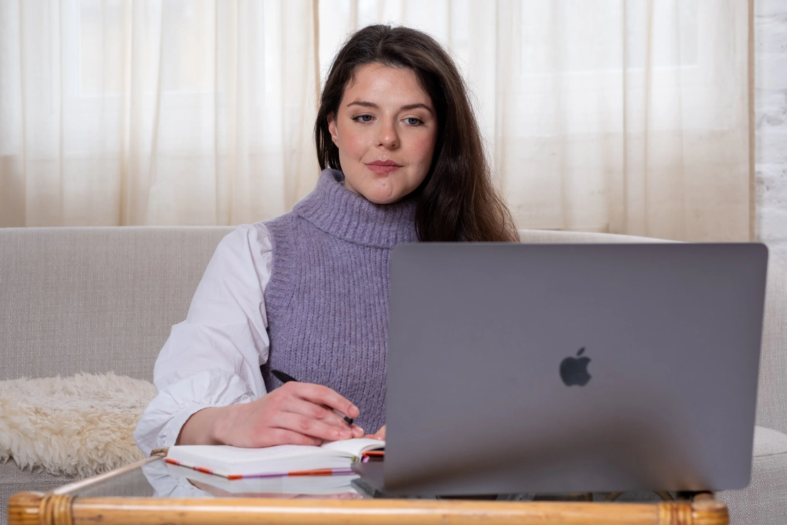 Bethan Stone, a woman with long brown hair and wearing a white shirt and purple sweater vest, sits at an Apple Macbook.