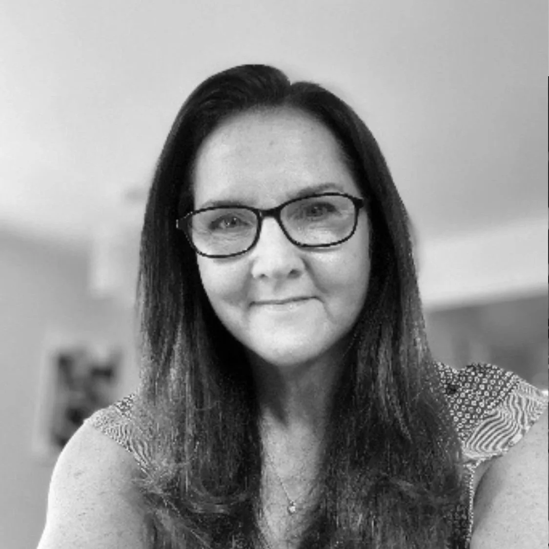 Black and white photo of a woman with long dark hair, glasses, lace top, smiling at the camera.