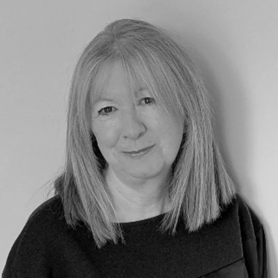 Black and white portrait of a woman with shoulder-length hair, smiling, wearing a dark top, standing against a plain wall.