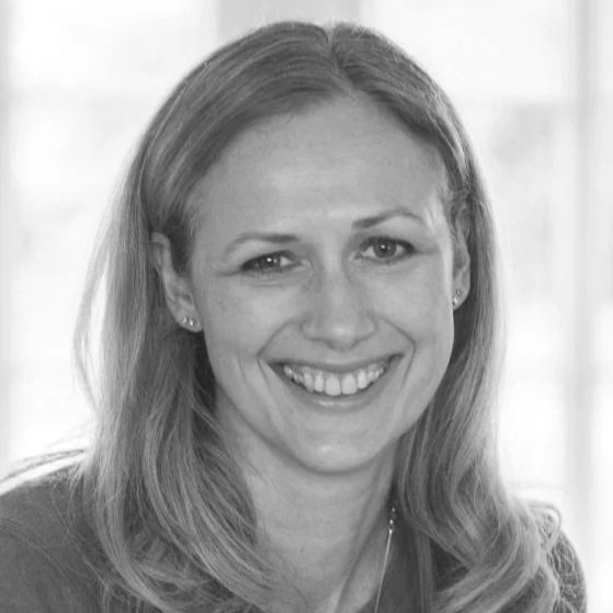 Black and white photo of a smiling woman with shoulder-length hair and earrings, indoors near a window.