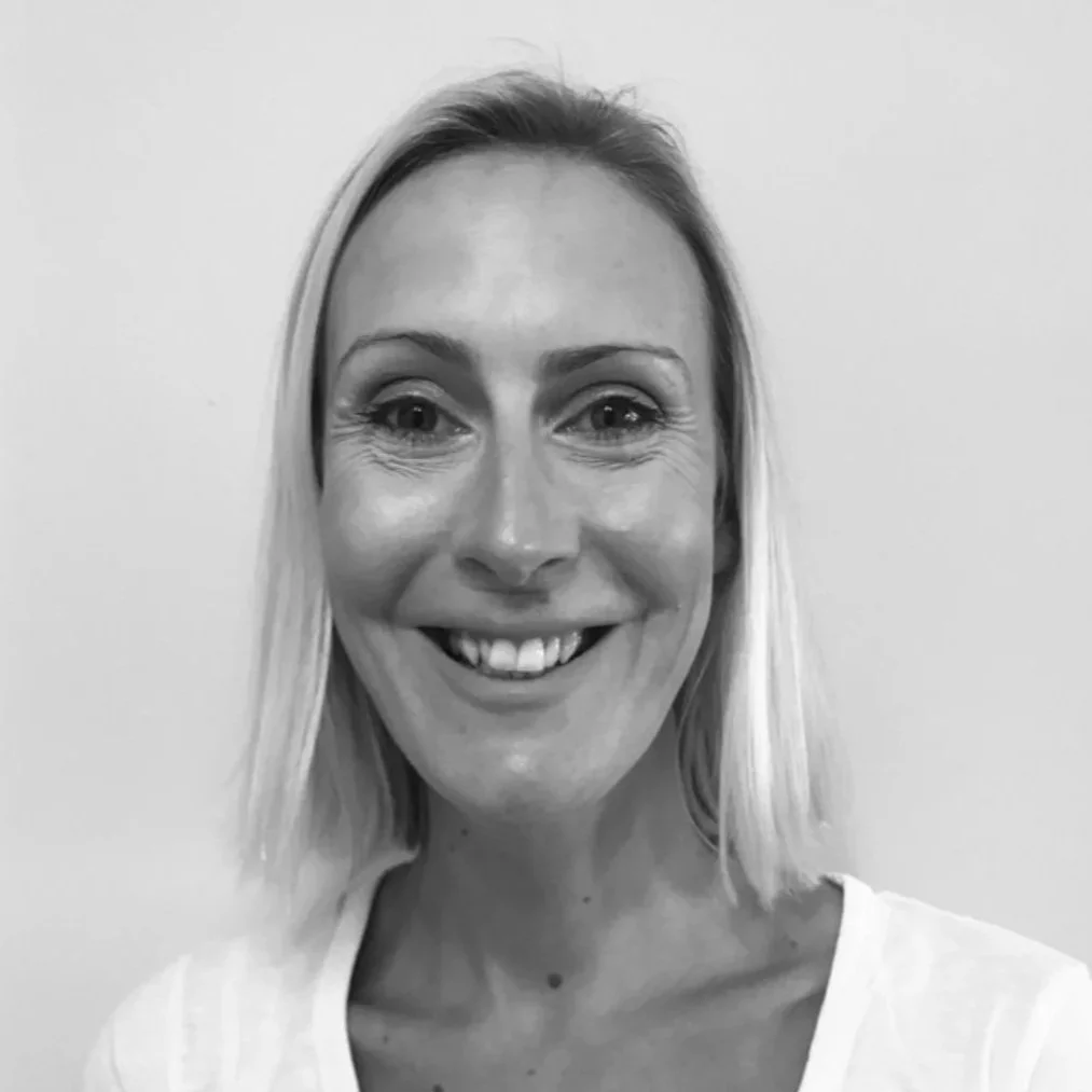 Black and white photo of a smiling woman with shoulder-length blonde hair, wearing a white top, standing against a plain wall.
