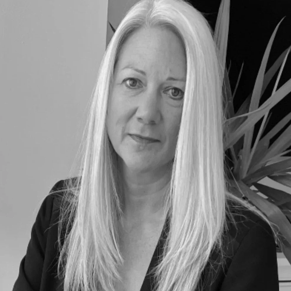 Black and white photo of a woman with long straight blonde hair, wearing a dark blazer, standing in front of a plant with long, pointed leaves.