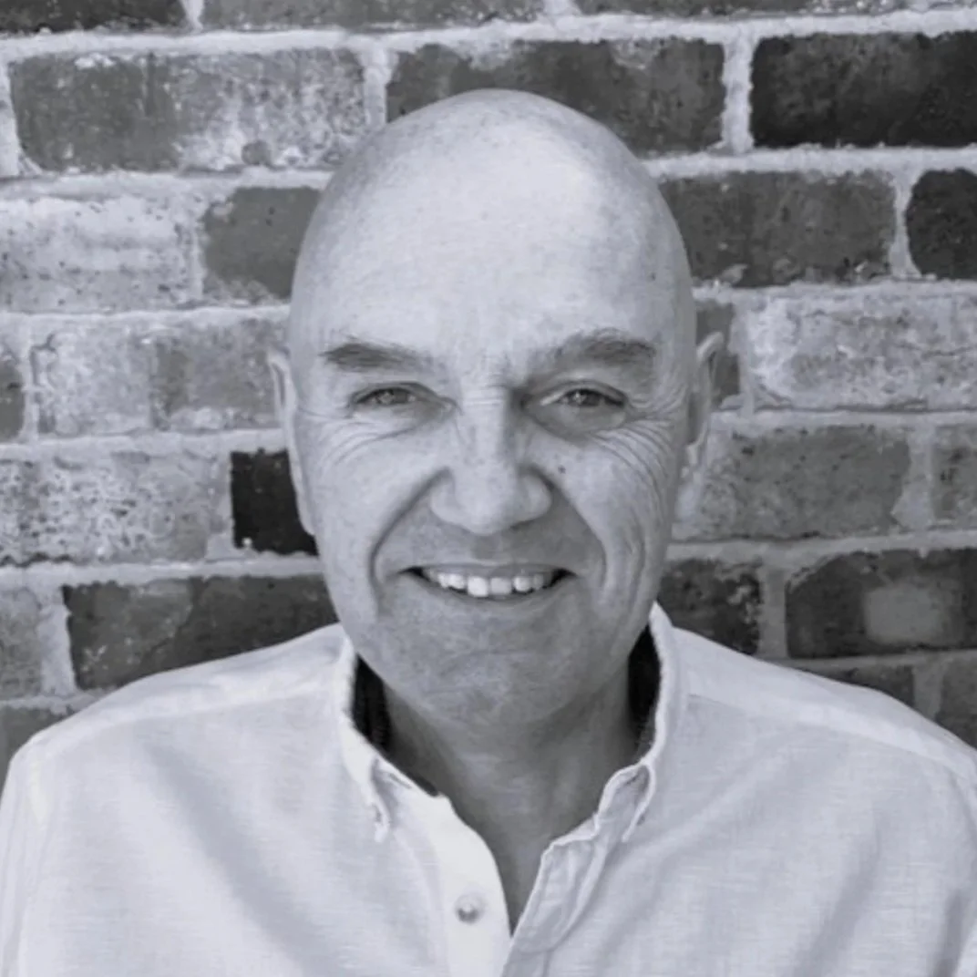 Black and white close-up of smiling bald man wearing a collared shirt, standing in front of a brick wall.