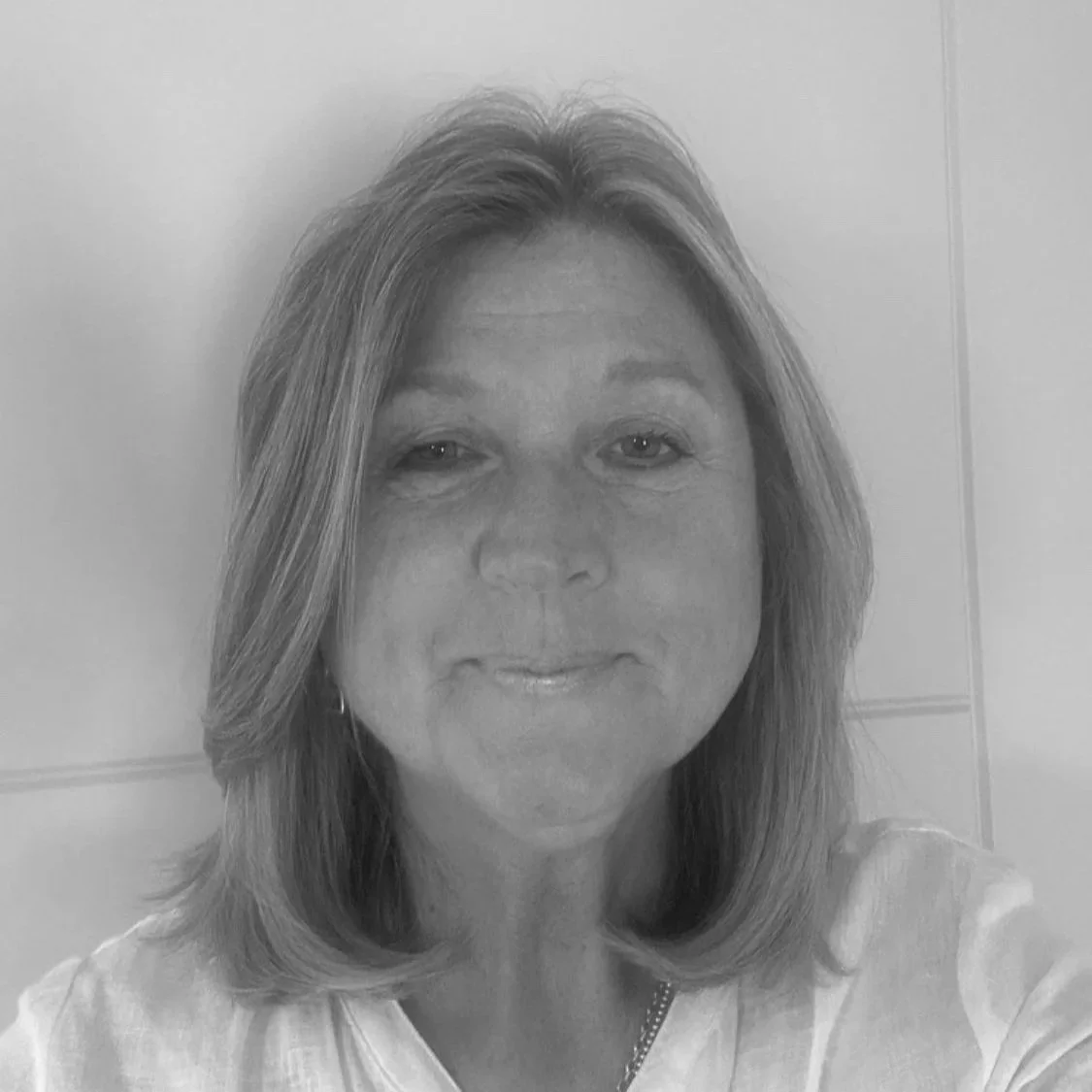 A woman with shoulder-length hair, smiling softly, wearing a light-colored top, in a room with plain walls.