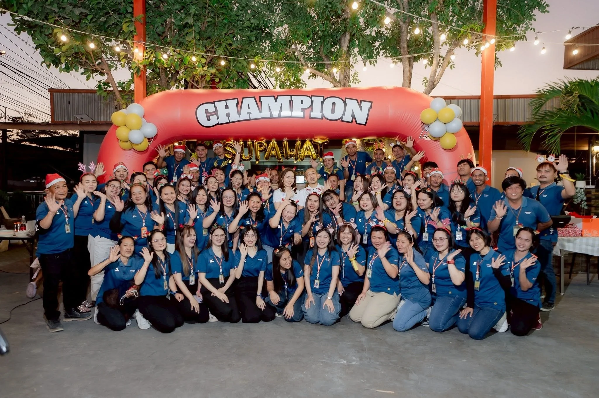 A group of approximately 50 people gathered outdoors, wearing blue shirts and holiday-themed accessories like Santa hats and reindeer antlers, under a large red inflatable arch with the word 'CHAMPION' and golden balloons spelling 'SUPALAT'. The scene is decorated with string lights and trees, suggesting a festive celebration or team event.