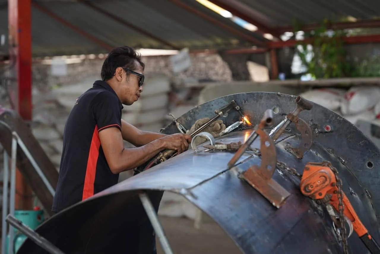 Man welding a large metal structure outdoors, wearing glasses and protective gloves.