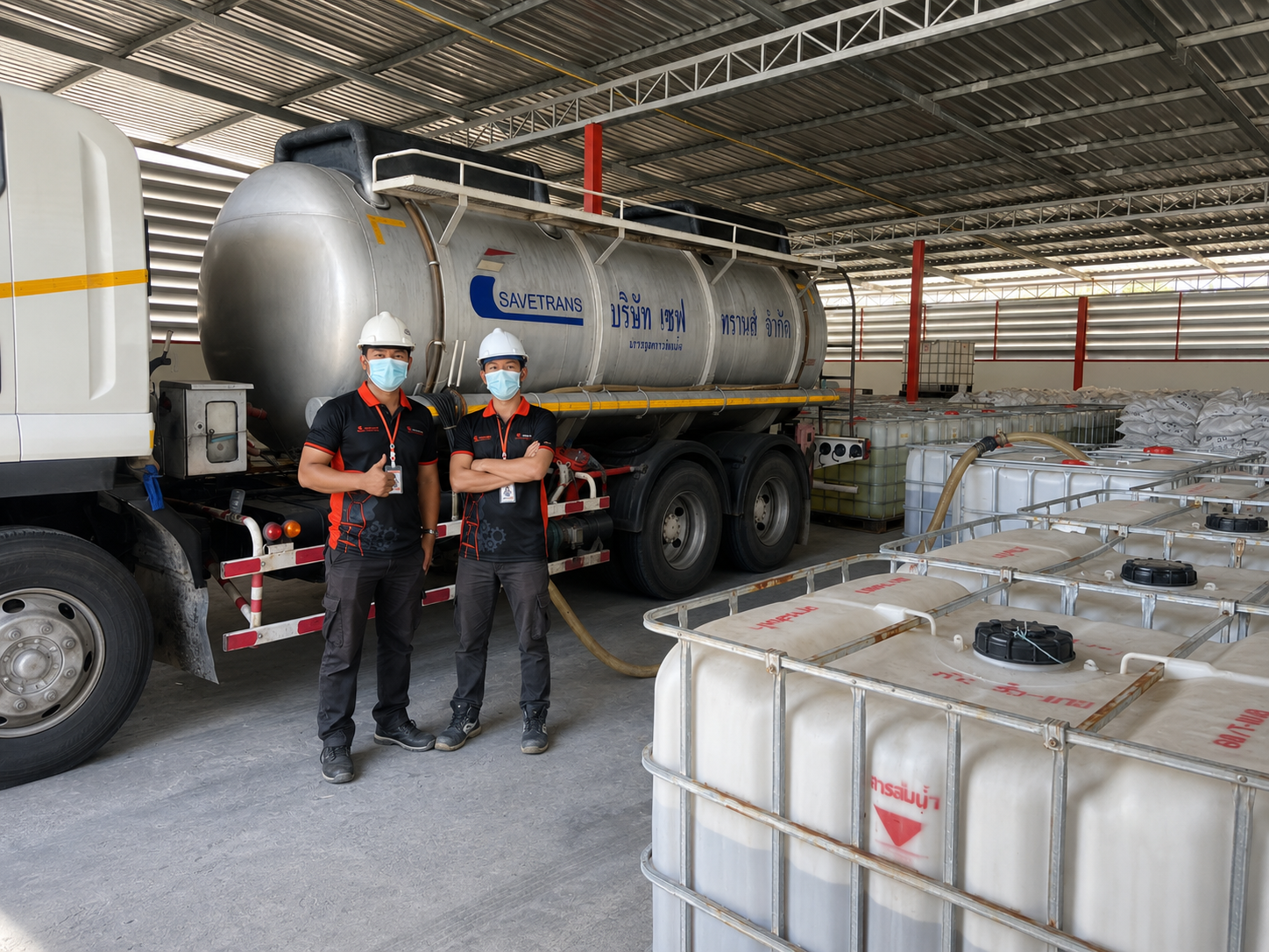 Two workers wearing safety helmets and face masks standing in front of a large tanker truck labeled 'SAVETRANS' inside an industrial facility. The truck is connected to large water tanks and equipment.