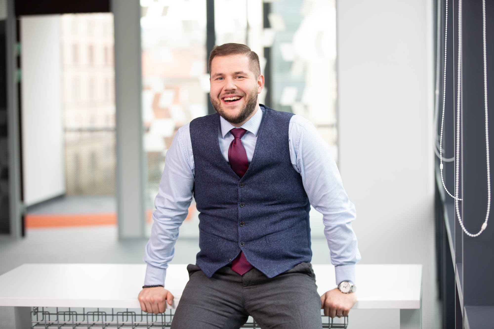 A man in a business suit with a vest, shirt, and tie sitting on a table in an office setting, smiling at the camera.