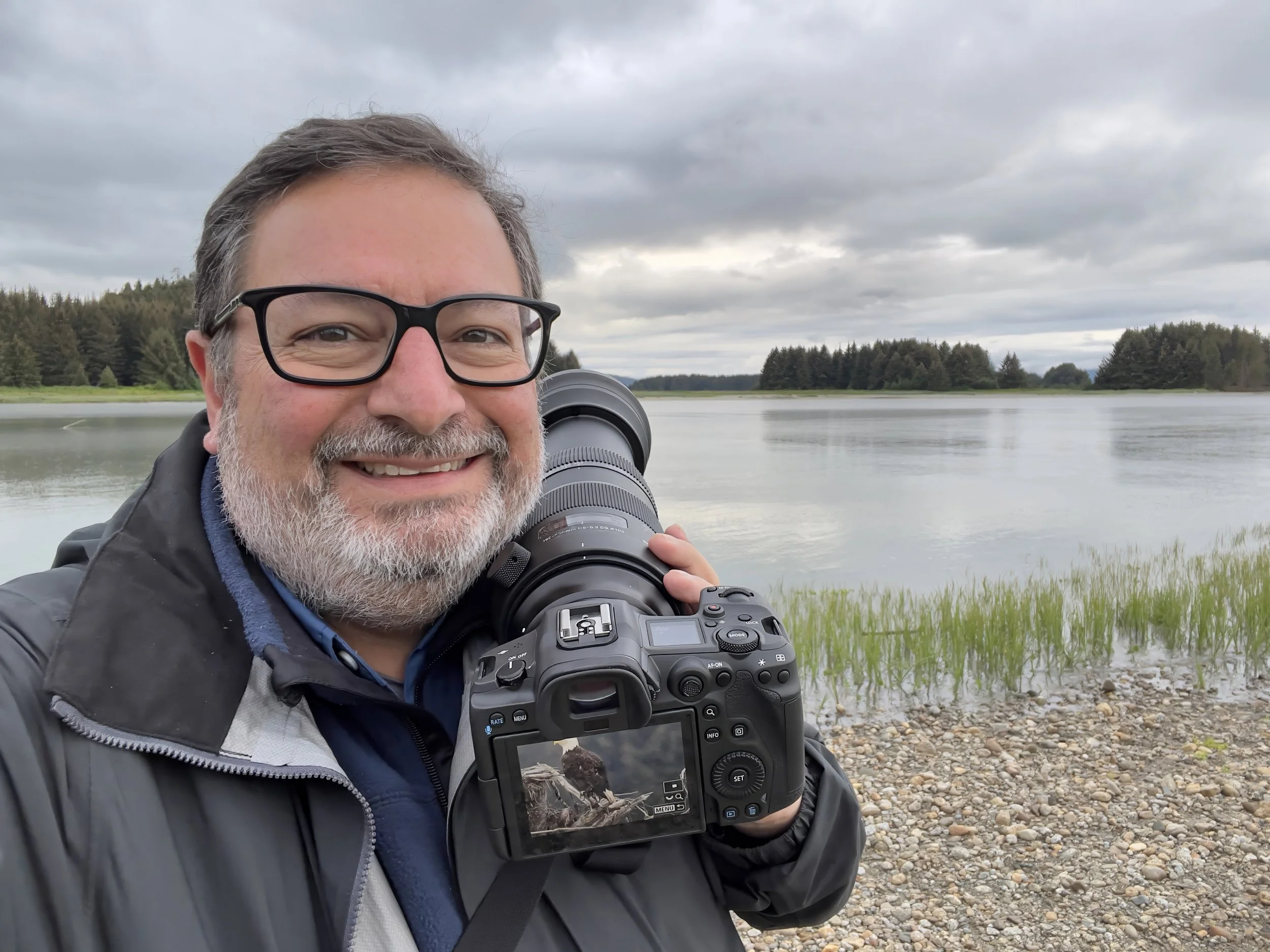 A smiling man with glasses and a beard holding a camera outdoors near a river or lake with a cloudy sky and trees in the background.
