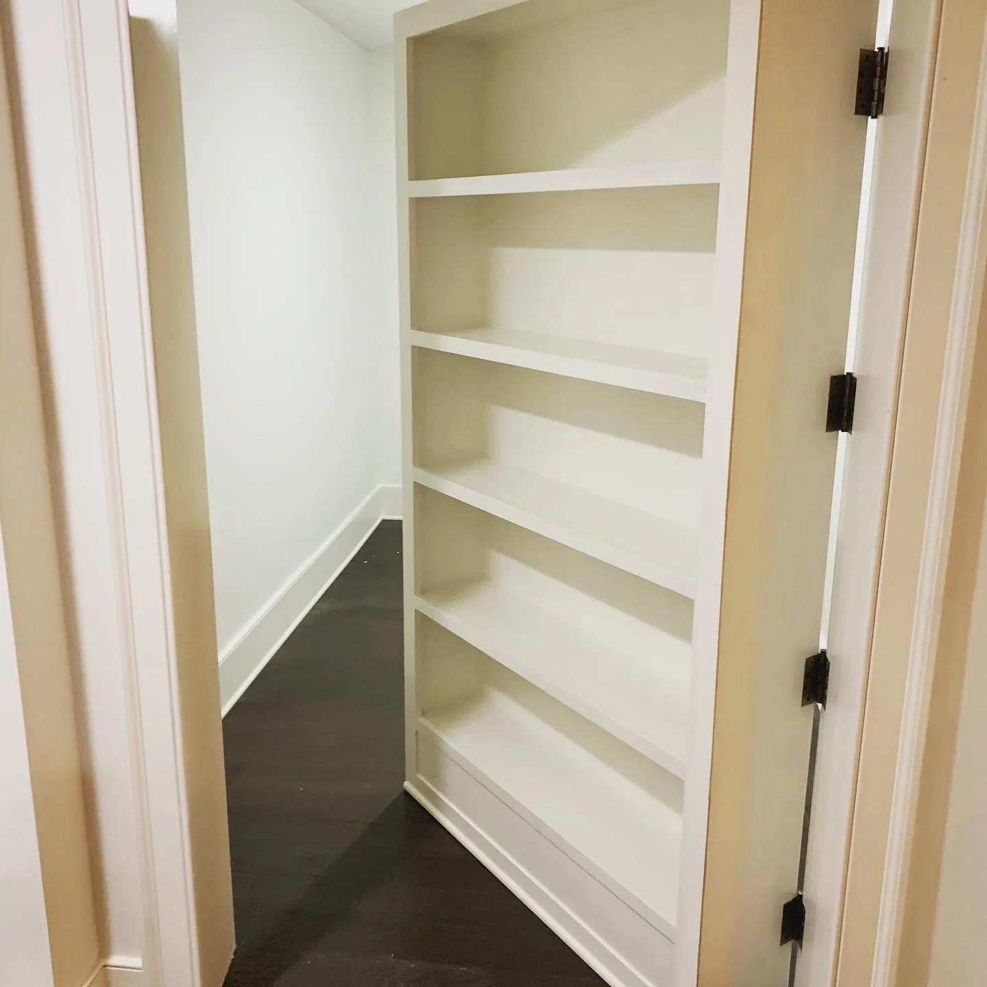 Custom built-in white wood shelving with a concealed hidden door shown in the open position, revealing the doorway while maintaining seamless integration through clean lines and matching paneling against neutral-toned walls.