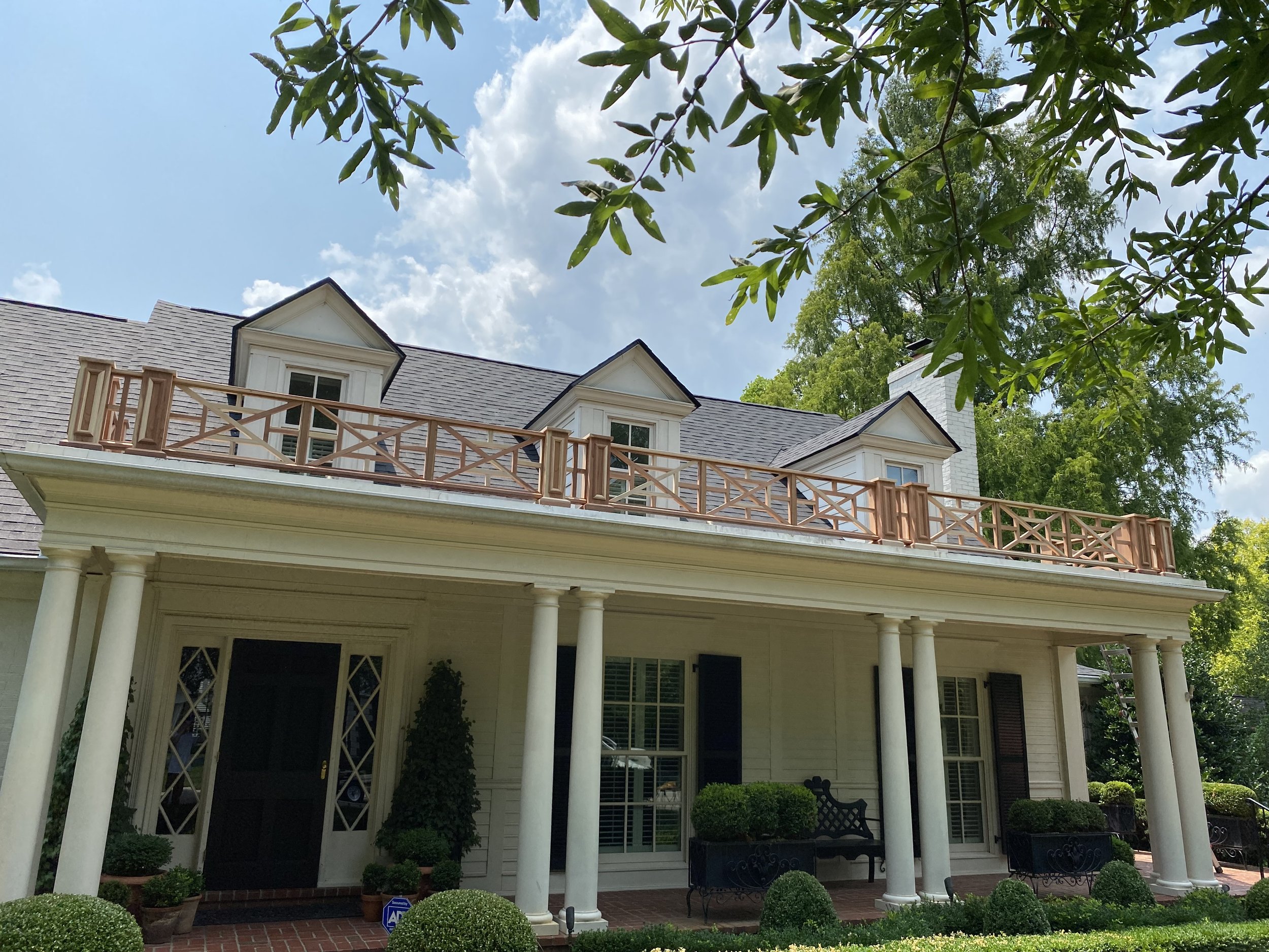 Rear view of a restored historic two-story home featuring a balcony with detailed railing, white columns, and a gray shingled roof, highlighting preserved architectural elements set against greenery and mature trees.