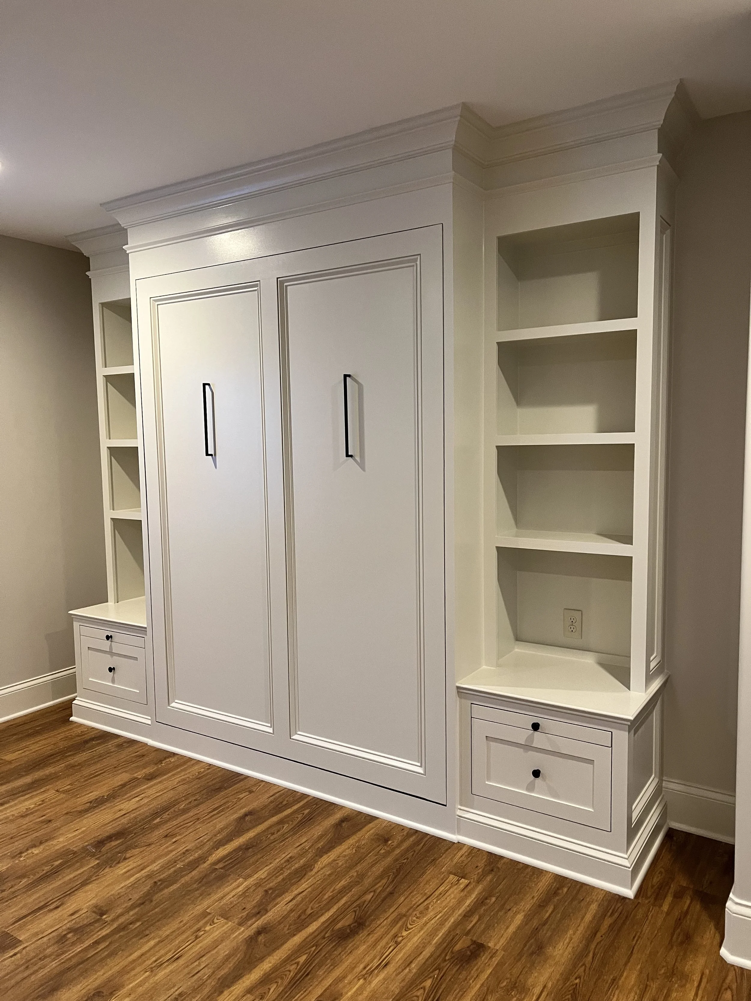 A built-in white wardrobe and shelving unit with black handles, situated against a wall with wood flooring.