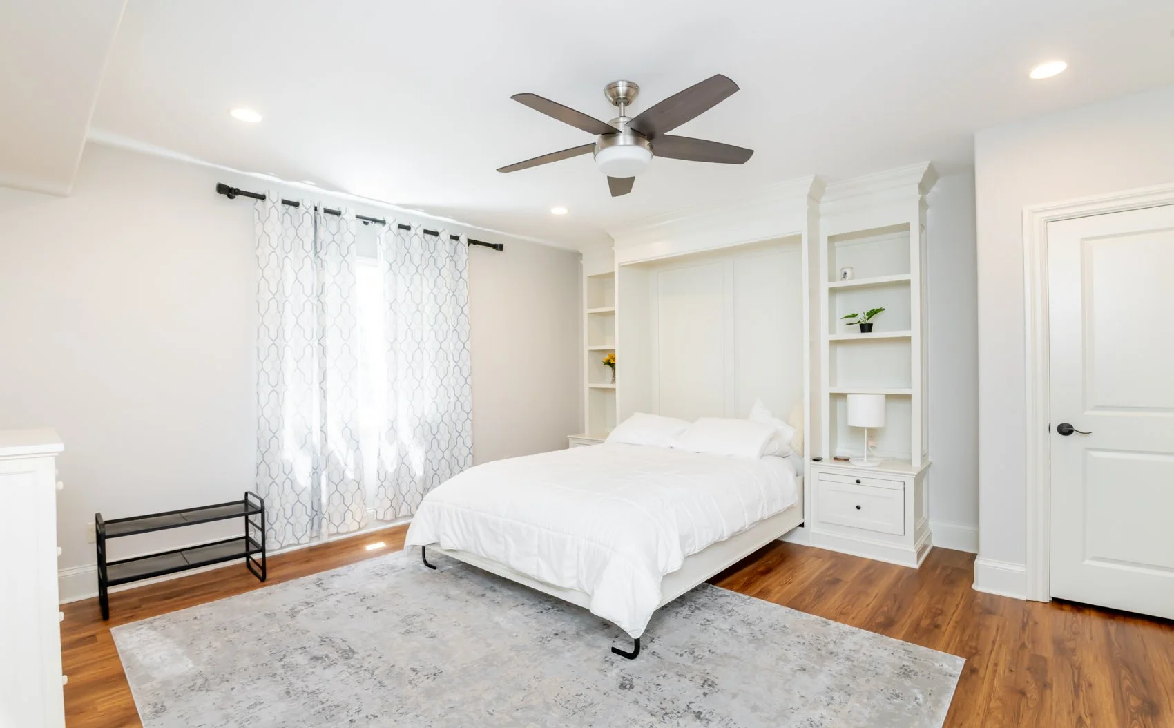 Bright bedroom with white walls, white bed, ceiling fan, hardwood floors, and built-in bookshelves with decorative items and a lamp.