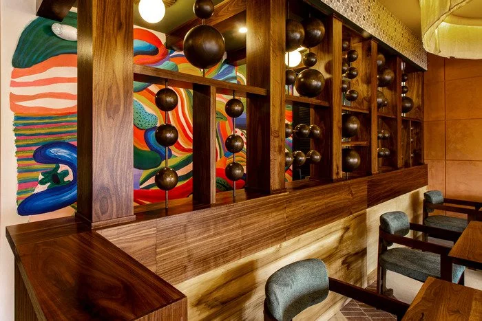 Gustavo's Mexican Grill - Louisville KY Interior of a restaurant with decorative wooden dividers featuring spherical ornaments, colorful abstract mural in the background, and tables with gray chairs.