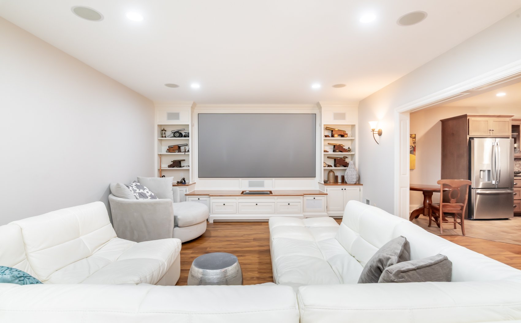 Built-in white wood entertainment center with integrated shelving and a recessed projector screen, showcasing clean architectural lines and decorative accents, with an open doorway revealing additional white wood cabinetry extending into the kitchen.