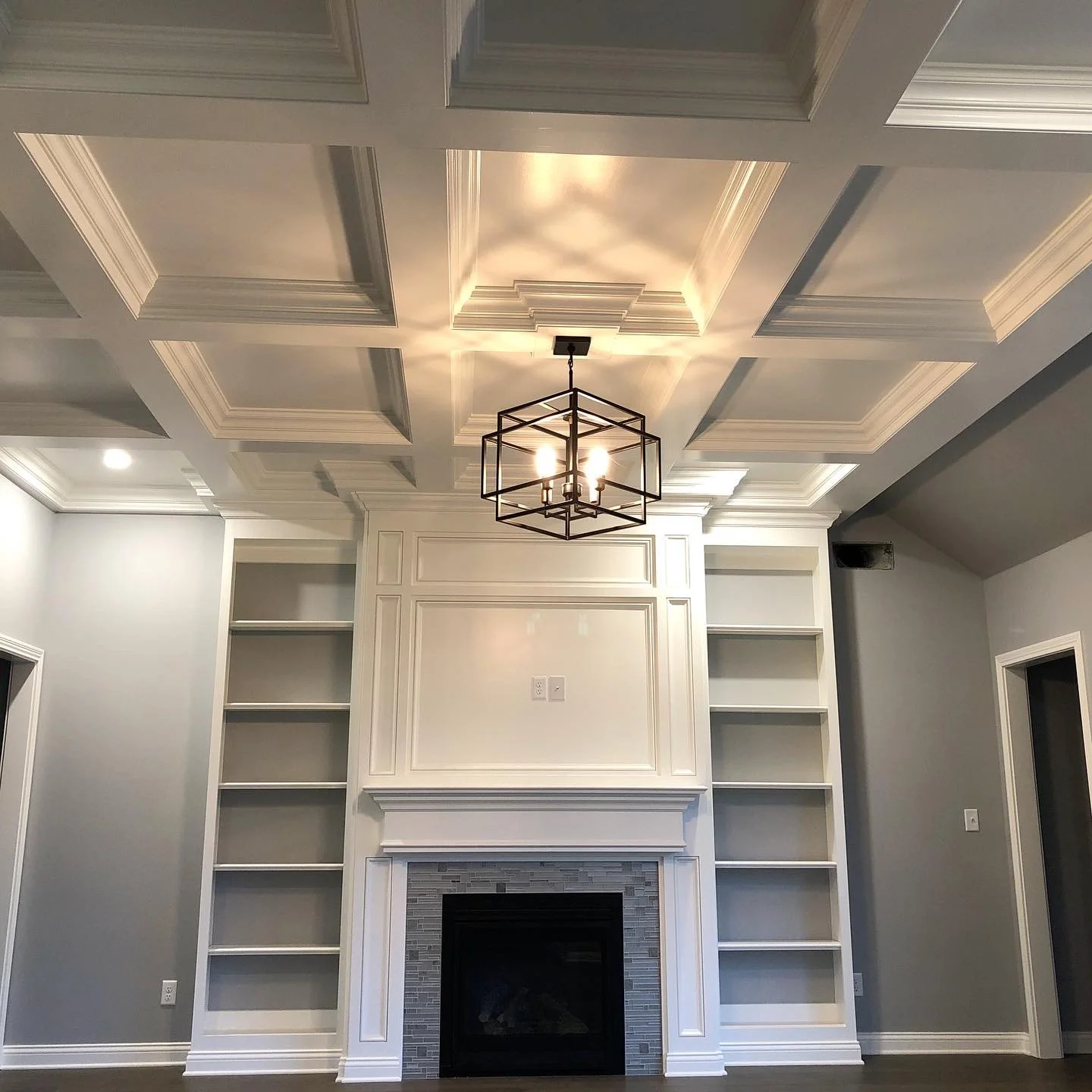Living room showcasing white wood built-in entertainment center and fireplace surround, complemented by a detailed coffered wood ceiling, gray walls, and clean architectural lines throughout the space.