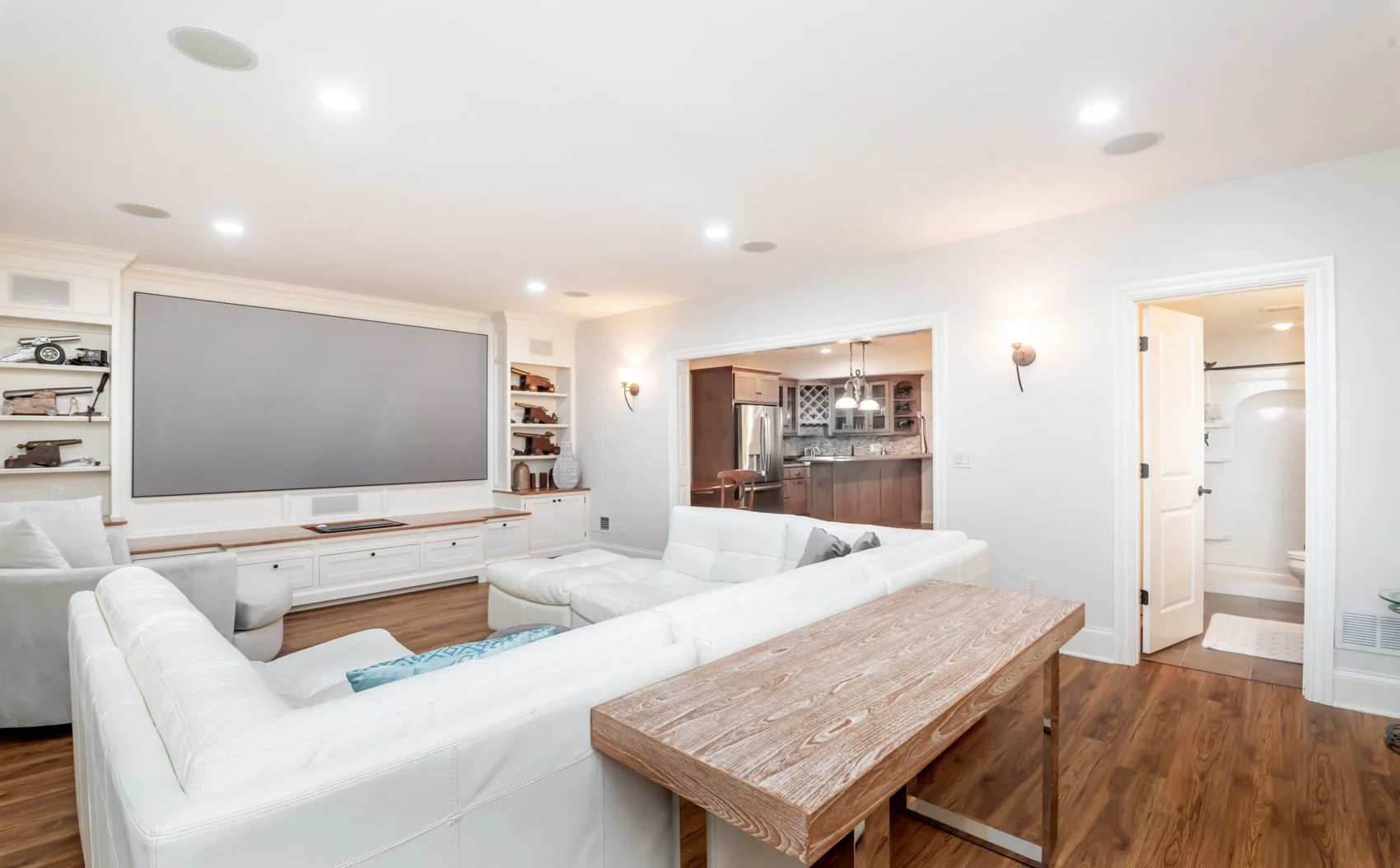 Living room featuring a custom-built wood entertainment center with integrated shelving and a recessed projector screen, showcasing clean craftsmanship and architectural wood detailing that anchors the space and opens toward the kitchen beyond.