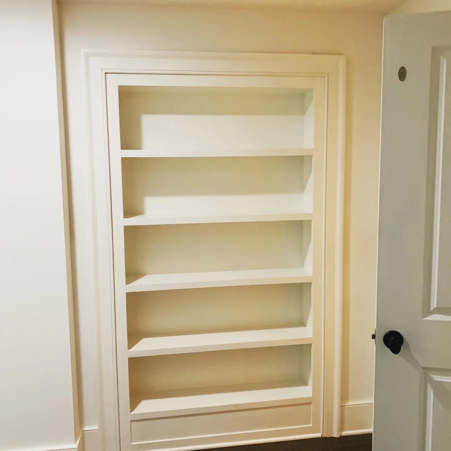 Custom built-in white wood shelving concealing a closed hidden door, designed to blend seamlessly into the wall with clean lines and integrated paneling against neutral-toned walls.