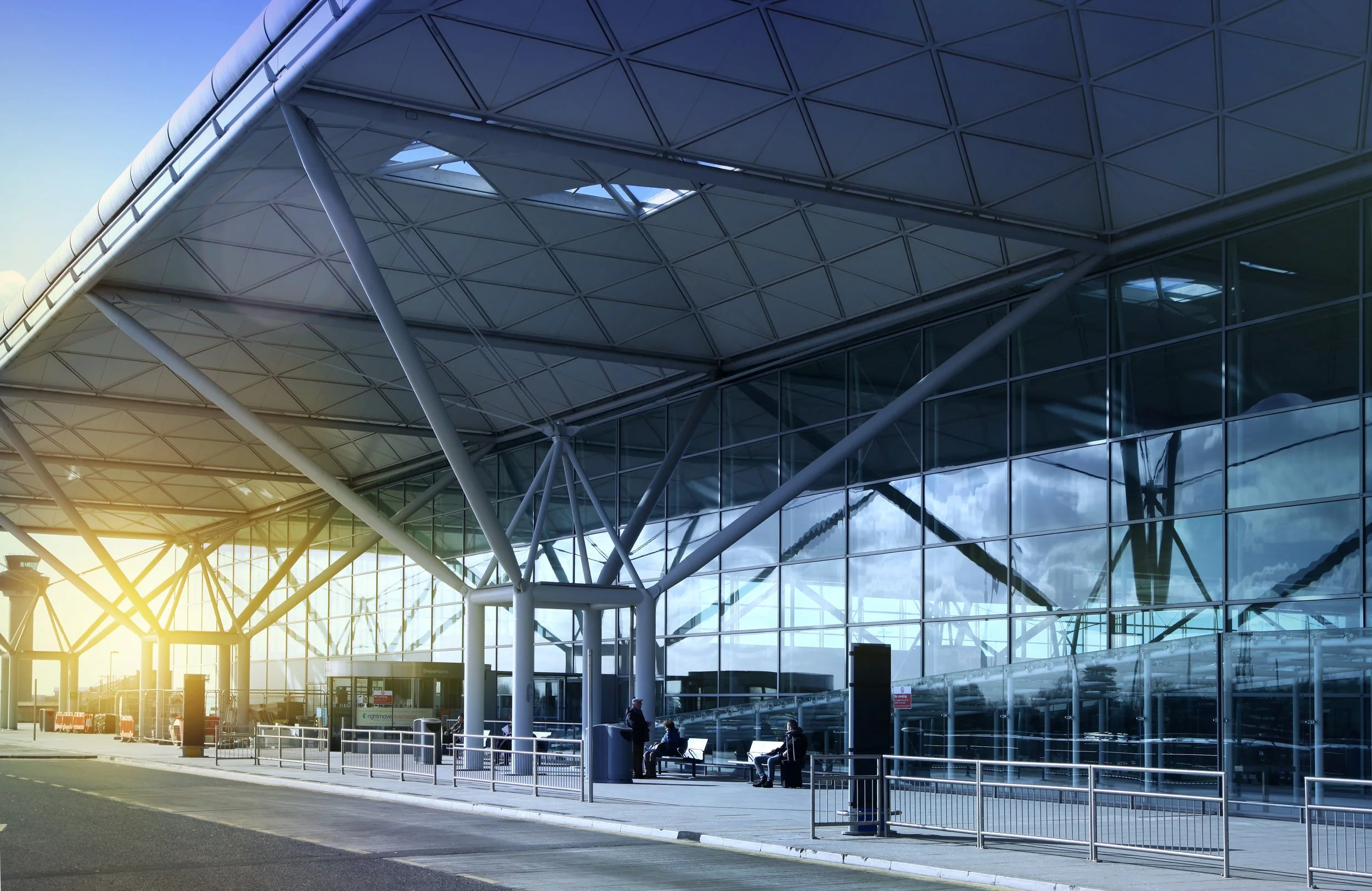 Modern airport terminal with glass walls and architectural metal beams, people sitting and waiting outside, with sunlight illuminating the scene.