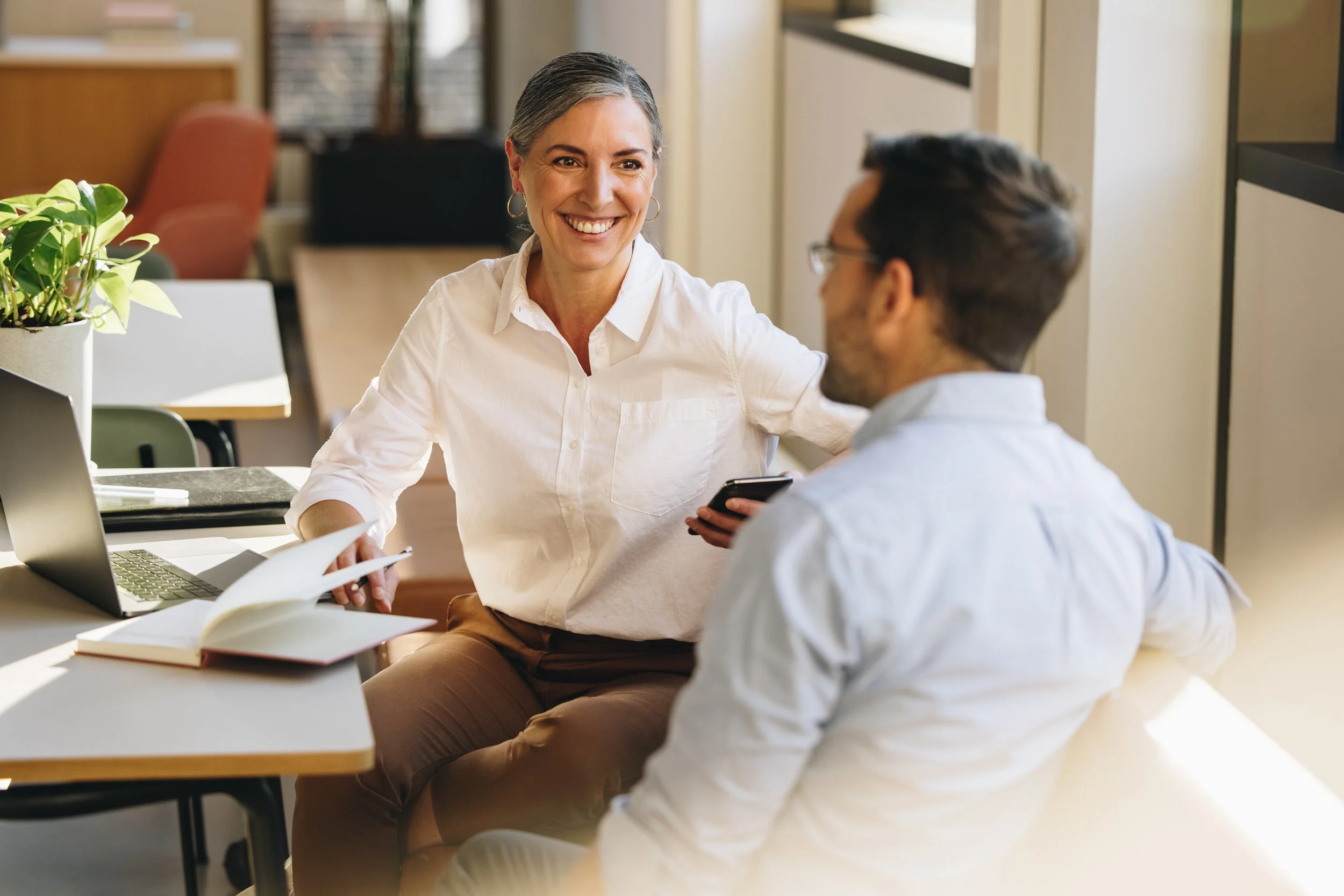 A woman with gray hair, wearing a white shirt, smiles while talking to a man with glasses and dark hair, sitting at a table in a well-lit office or meeting space with books, laptop, and a plant nearby.