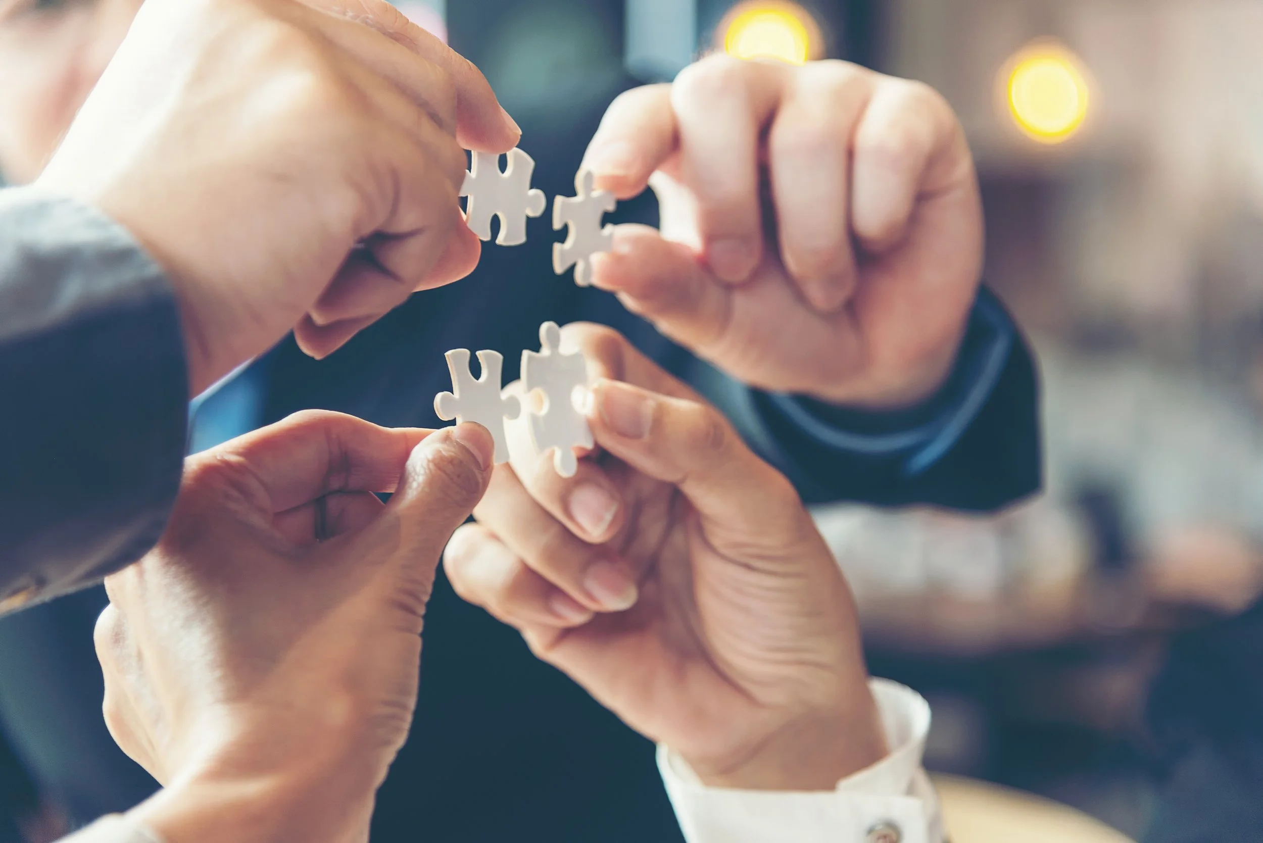 Multiple hands assembling white puzzle pieces, with a blurred background featuring warm yellow lights.