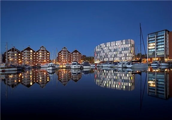 Boats docked at a marina with modern apartment buildings in the background at night.