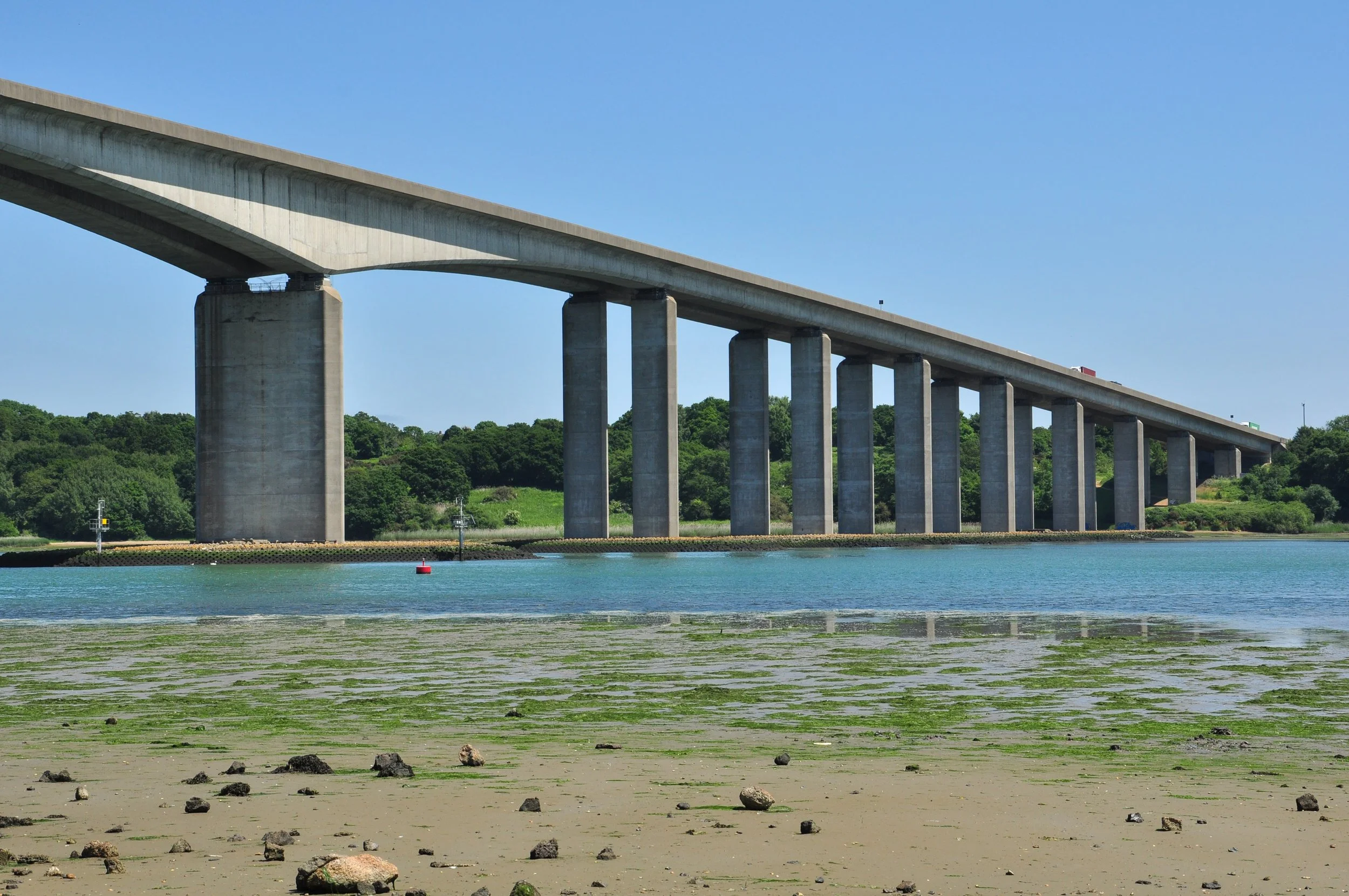 A concrete bridge extends across a river with a green shoreline and a clear blue sky.